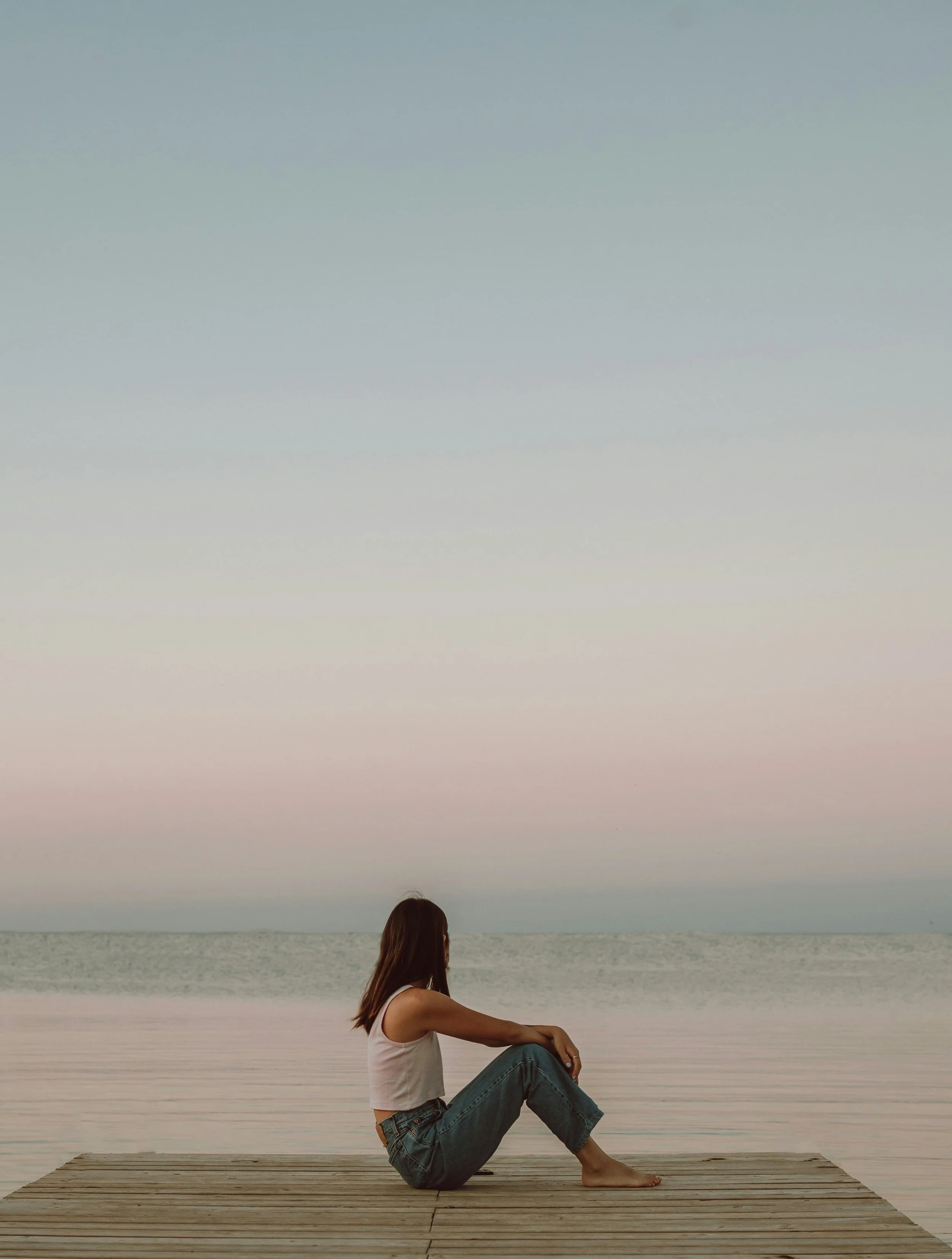 A woman sitting on a wooden dock by the water during sunset or dusk, with her back and side to the camera, wearing a white tank top and denim jeans.