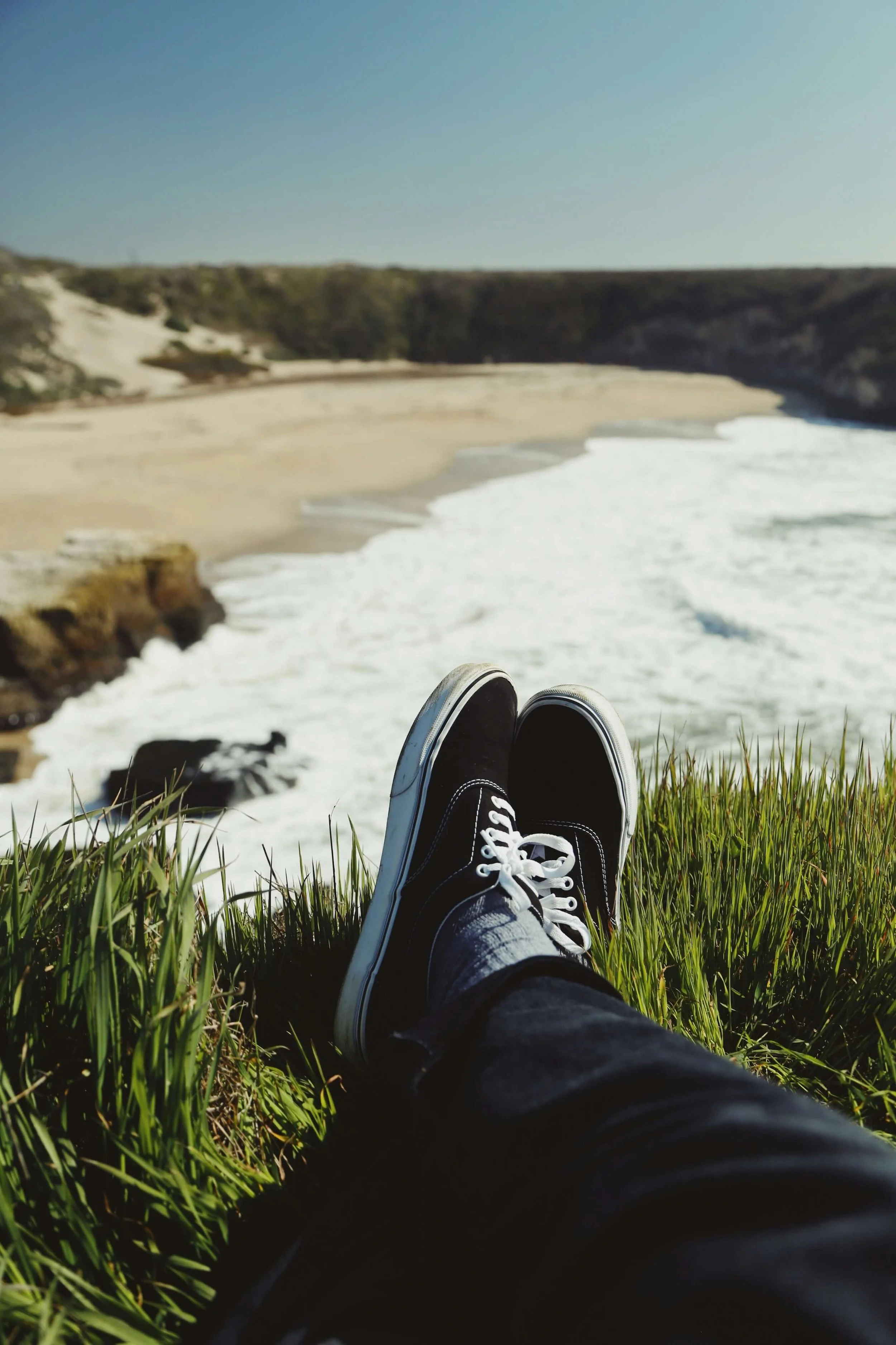 Person lying on grass, wearing black sneakers with white laces, overlooking a beach with waves and cliffs under a clear blue sky.