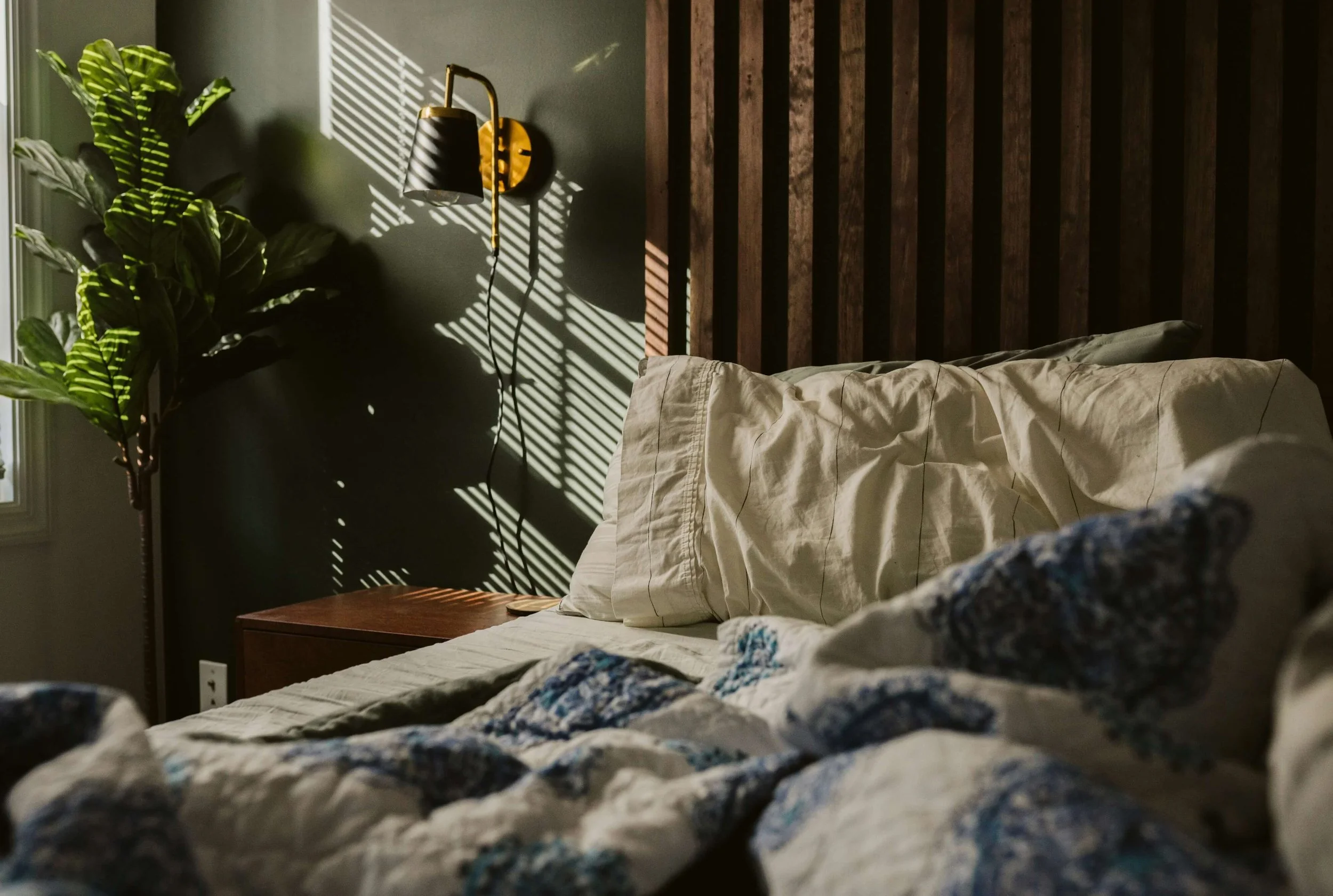 A bedroom with a bed, a wooden headboard, a nightstand, a wall-mounted lamp, a potted plant, and sunlight casting shadows through window blinds.