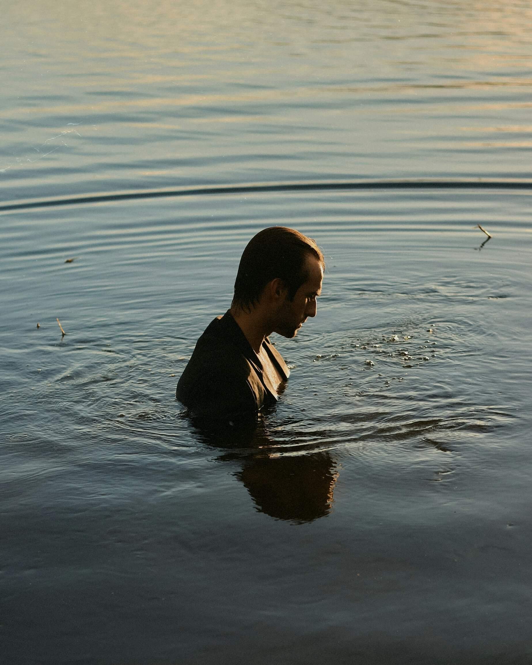 A man with dark hair, wearing a black suit, is standing in a body of water, with the water reaching up to his shoulders, and his reflection visible on the surface of the water.