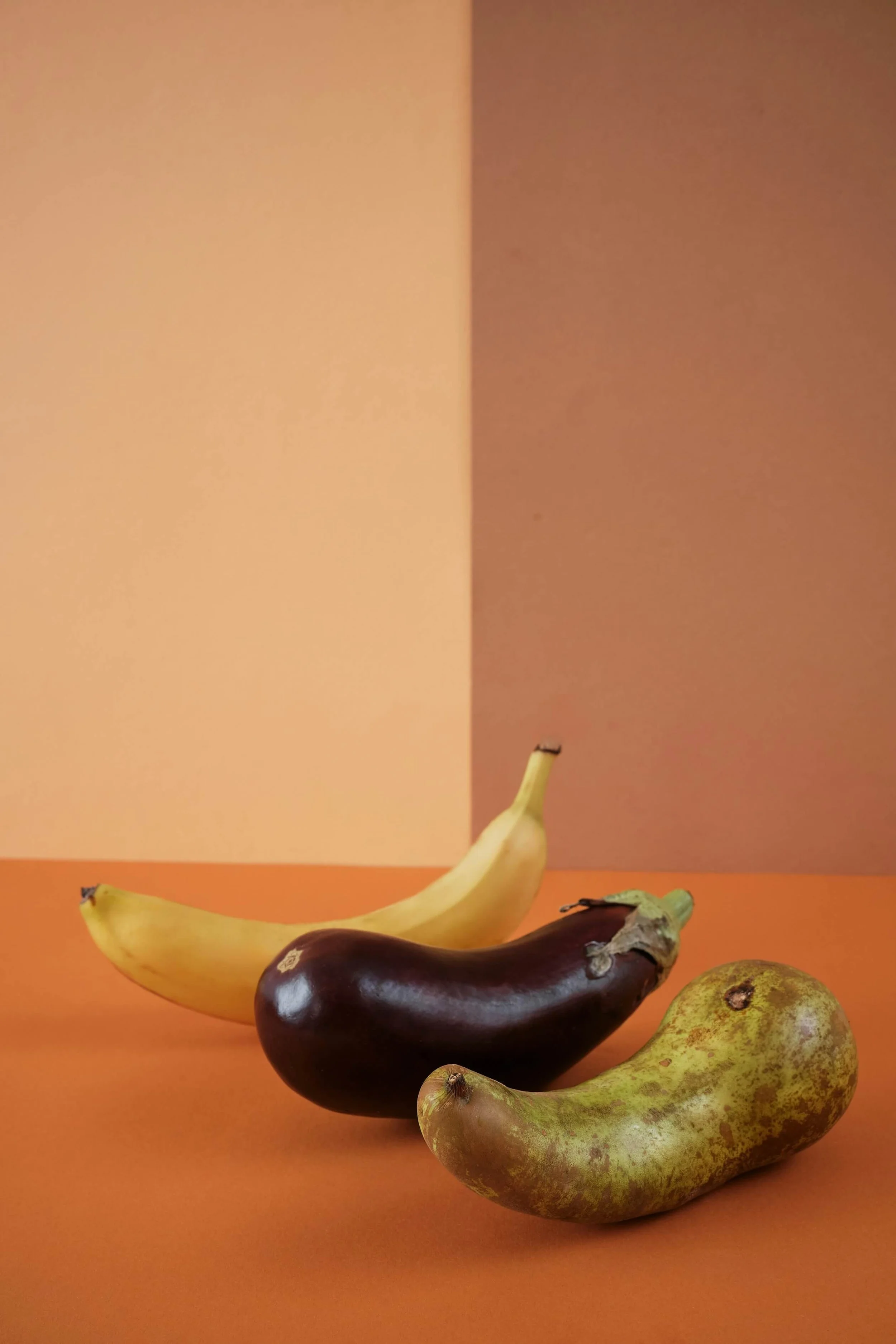 A still life of various bananas and pears on an orange surface with a two-tone background, featuring a yellow banana, a purple eggplant, a green pear, and a yellow pear.