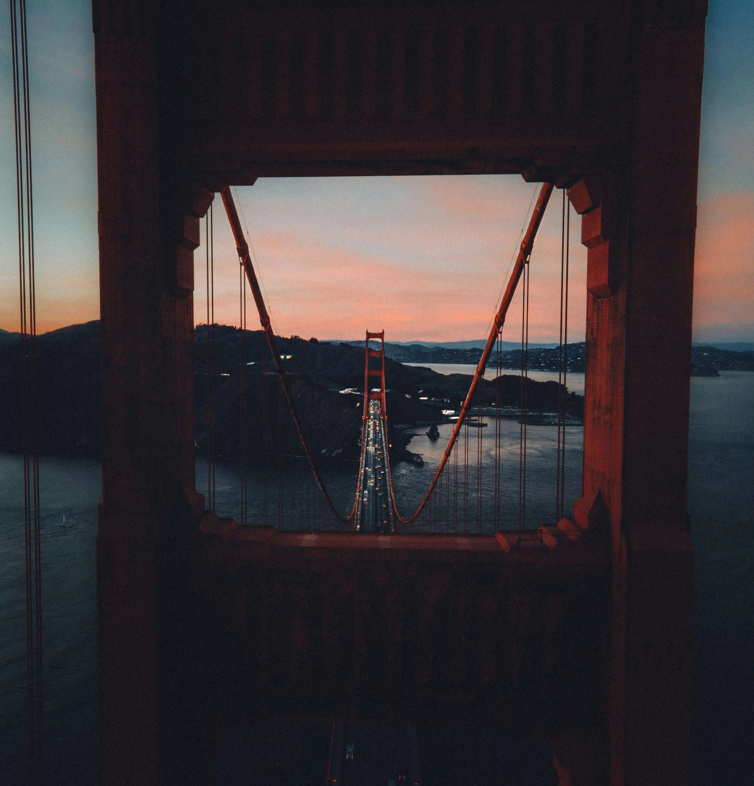 View of the Golden Gate bridge at sunset with tall support towers, cables, and a roadway. The sky displays a gradient of colors from pink to blue, and mountains are visible in the background.