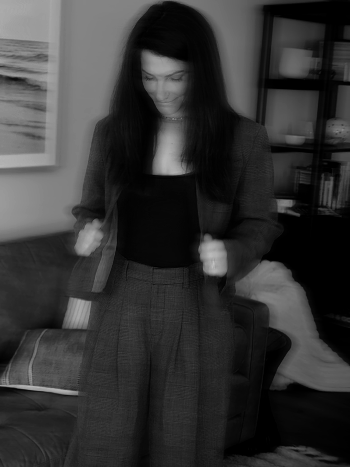 A woman with dark hair, dressed in a checkered suit, standing indoors in front of a bookshelf and a painting, looking down and smiling.