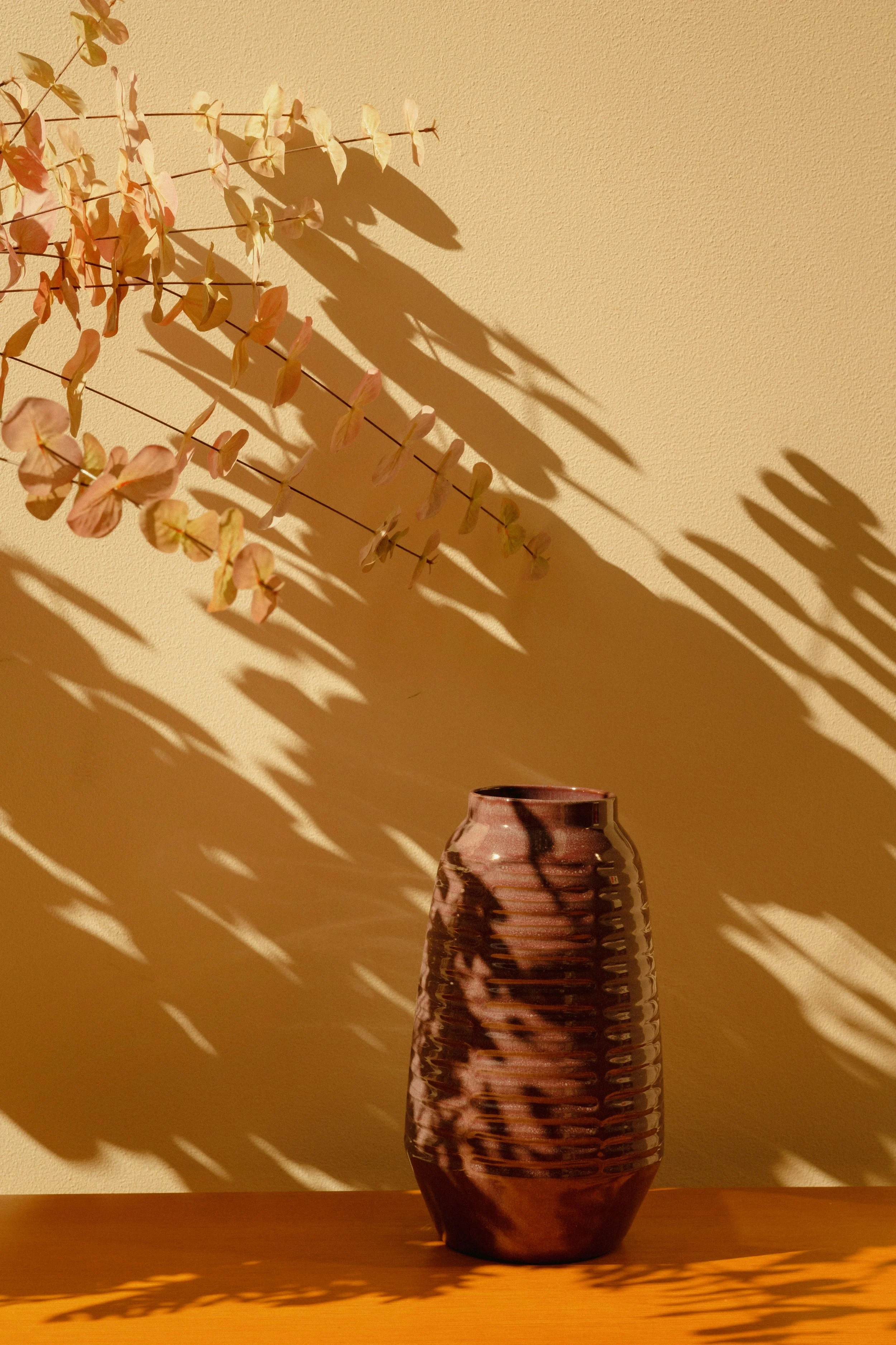 A tall, brown, textured glass vase on a wooden surface with shadow patterns cast on a beige wall behind it.