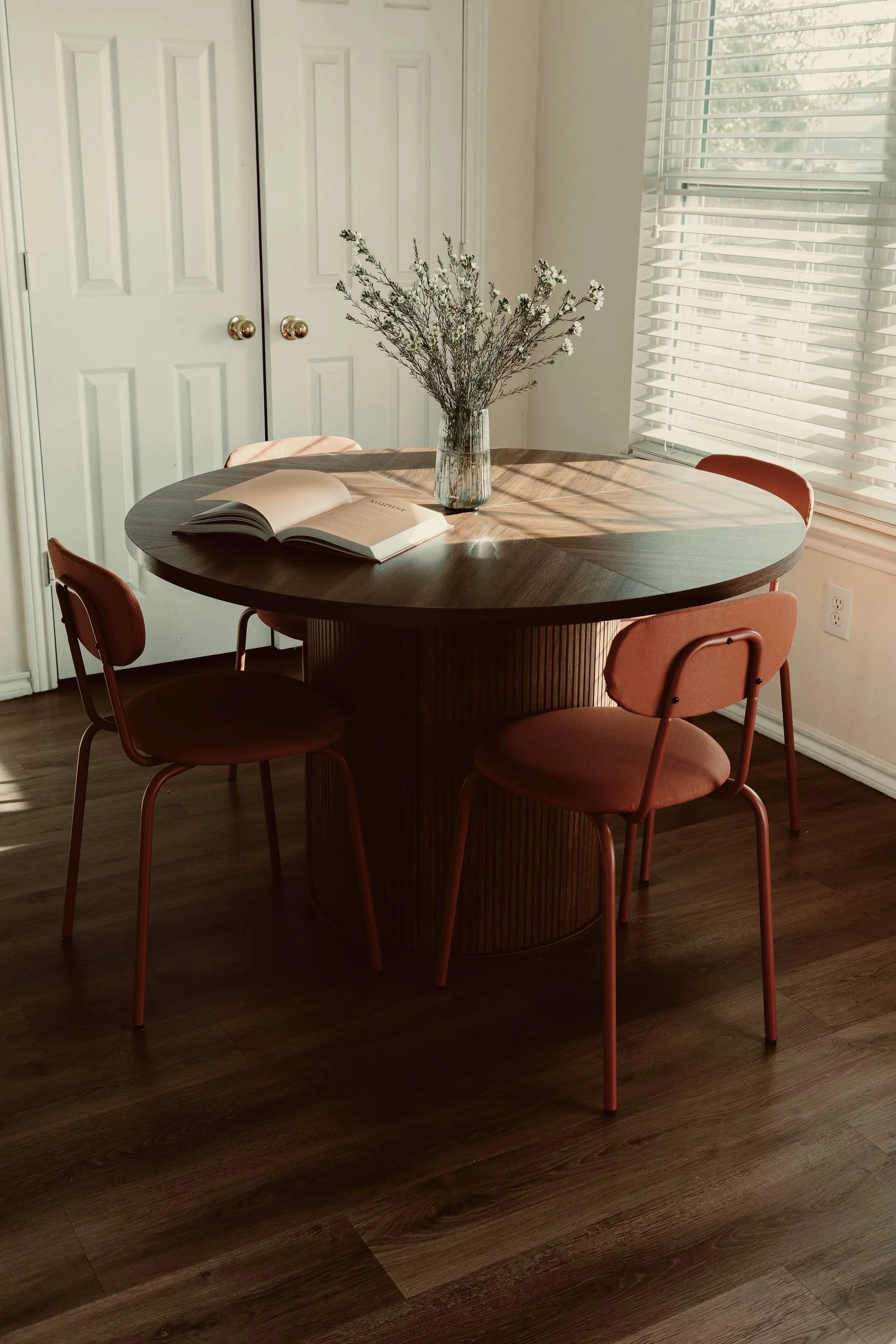 A round wooden dining table with a glass vase of white flowers, an open book, and pink chairs in a sunlit room with white blinds and closed closet doors.