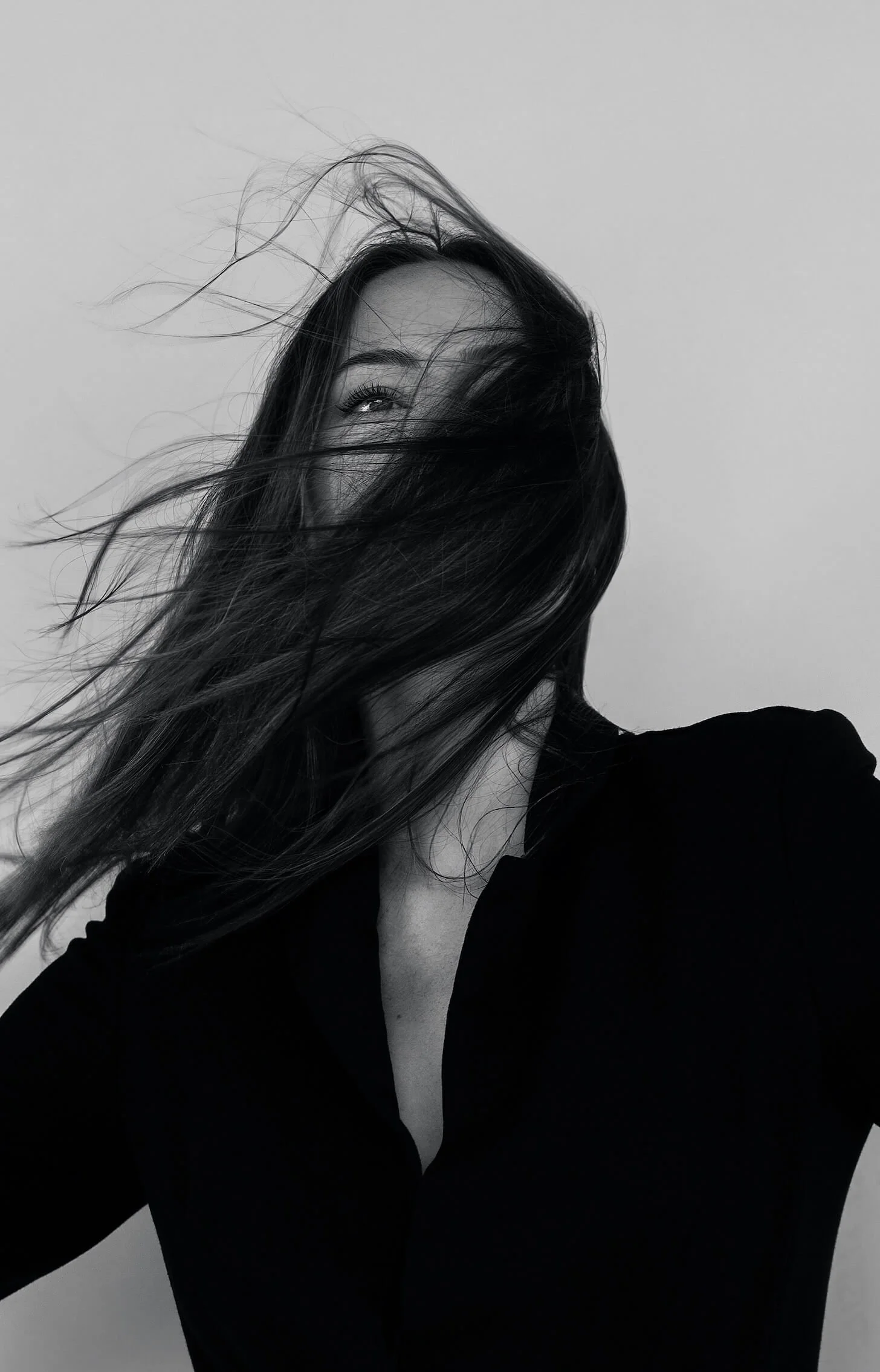 Black and white photo of a woman with long hair blowing in the wind, wearing a dark top, looking up.