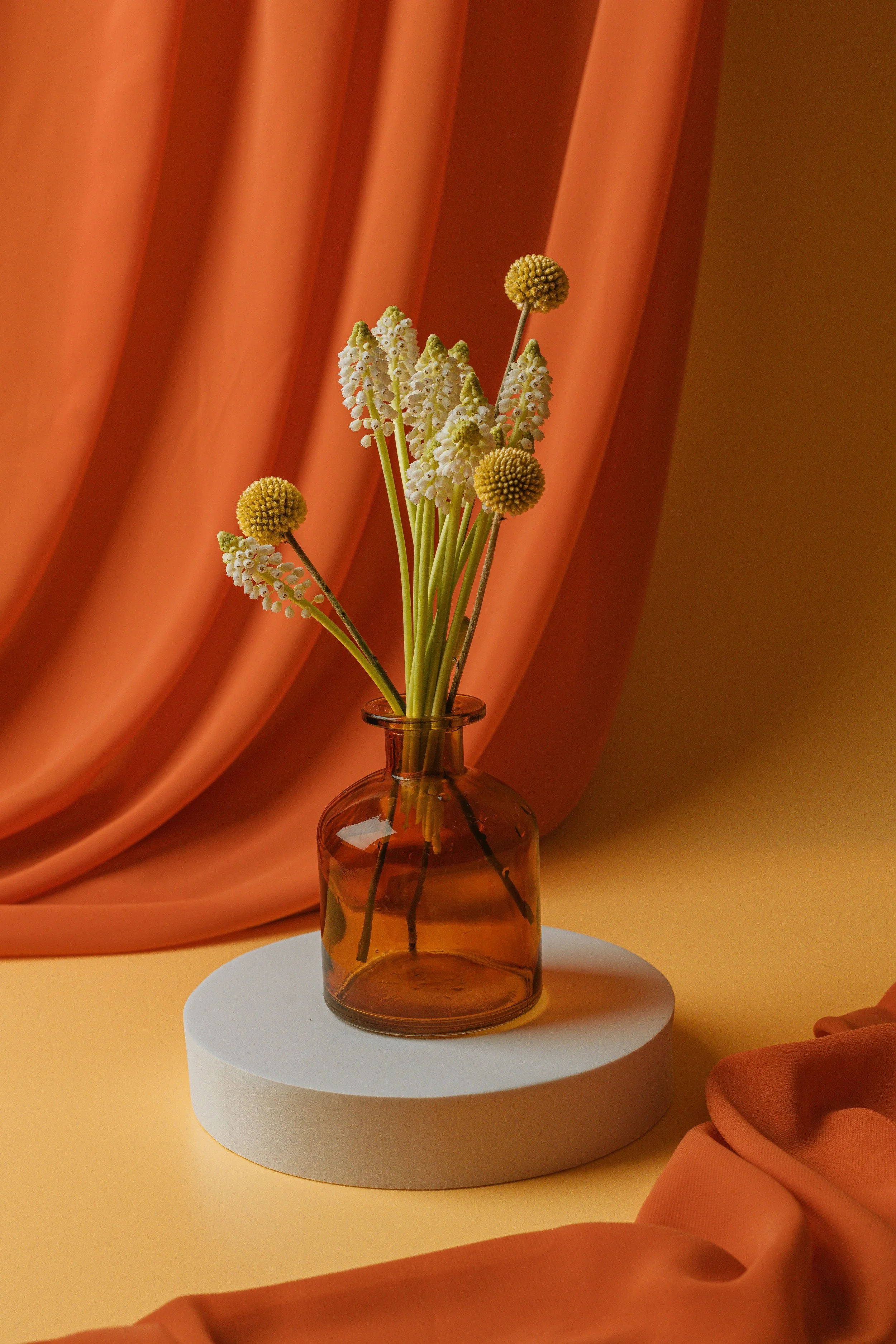 A bouquet of white and yellow flowers in a small amber glass vase, placed on a round white pedestal, with a warm yellow background and orange draped fabric.