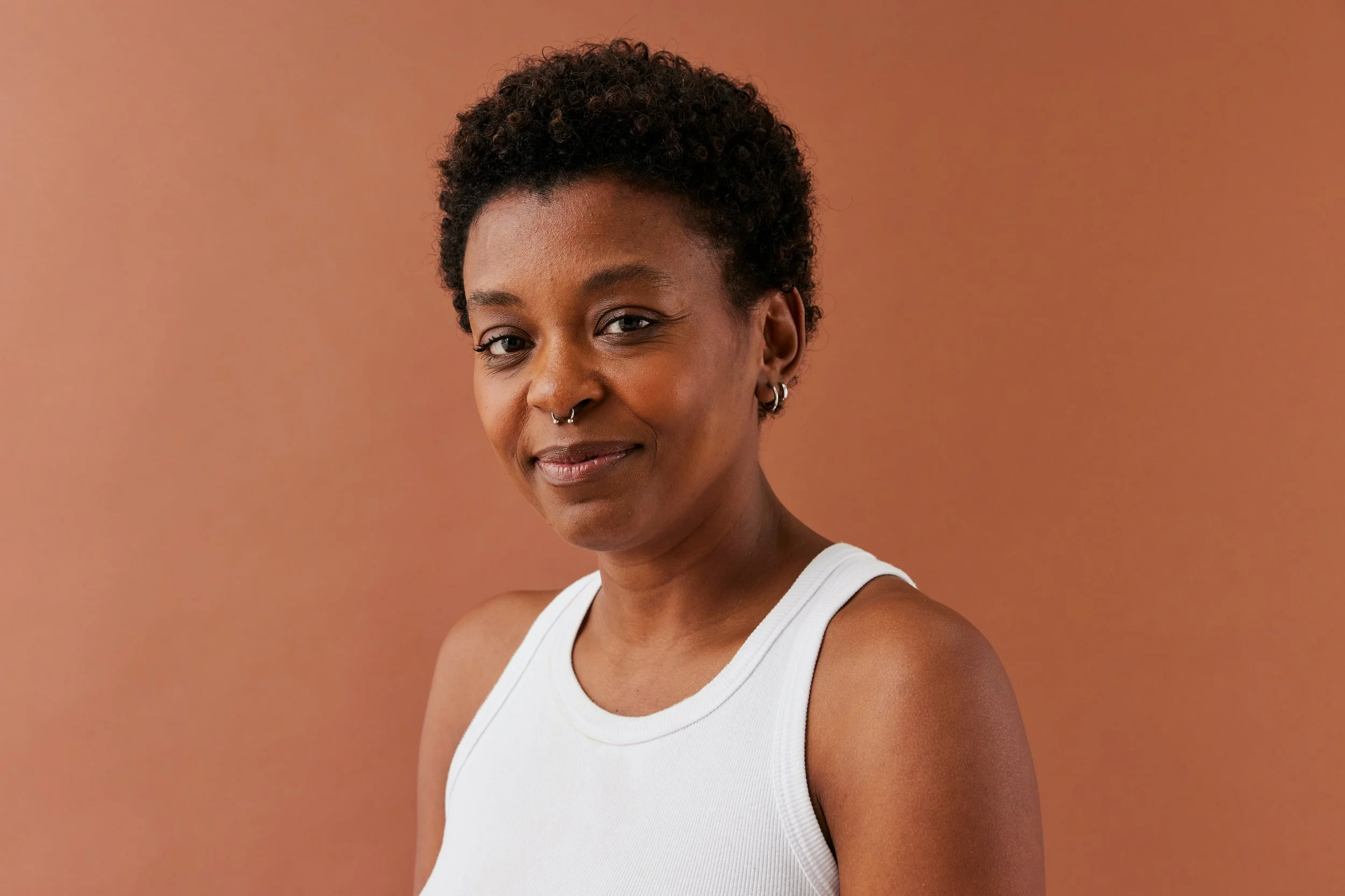 Close-up of a woman with short curly hair, wearing a white tank top, smiling slightly, with a neutral brown background.