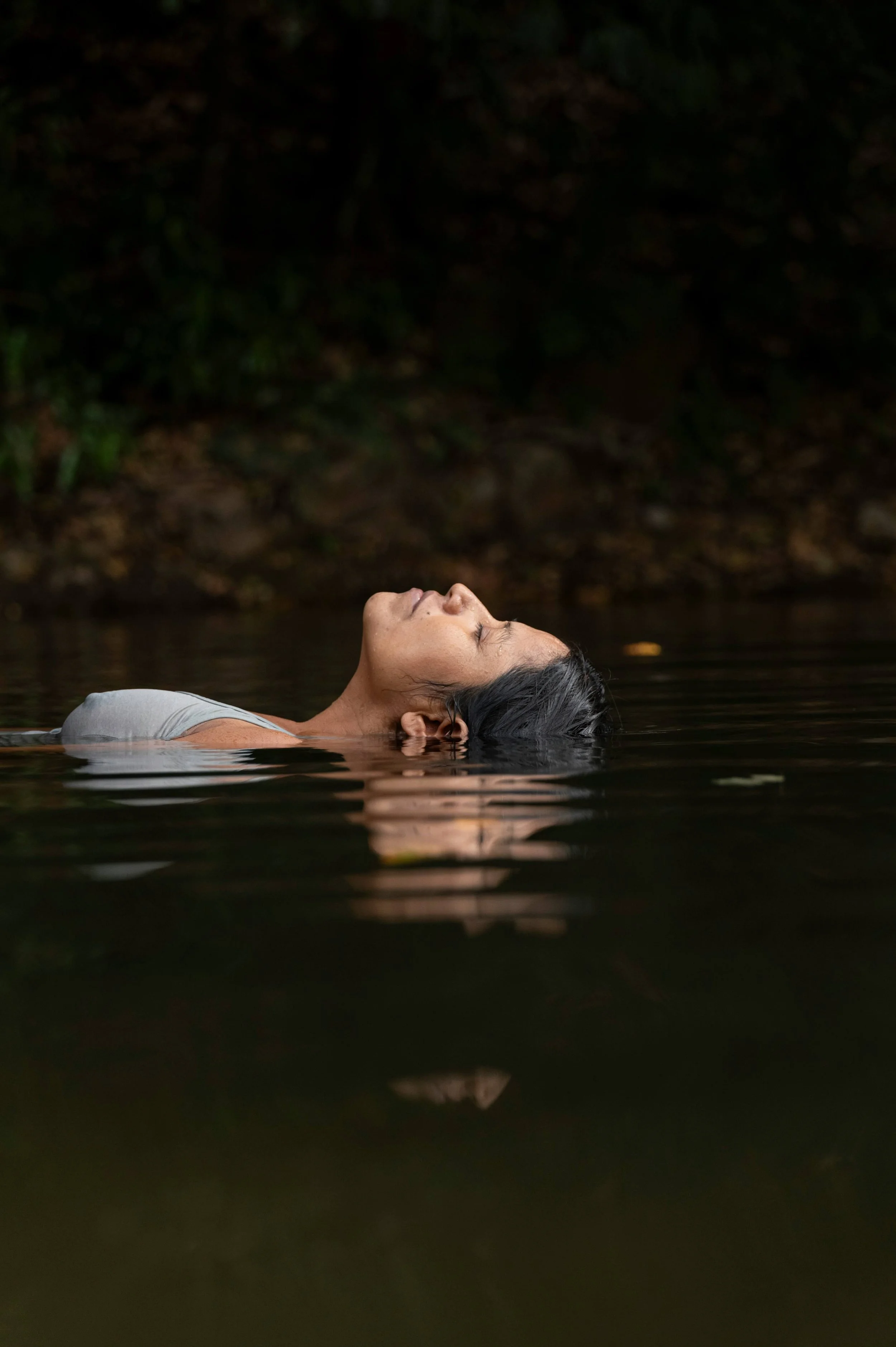 A woman with dark hair relaxes in water outdoors with her eyes closed, surrounded by dark foliage.