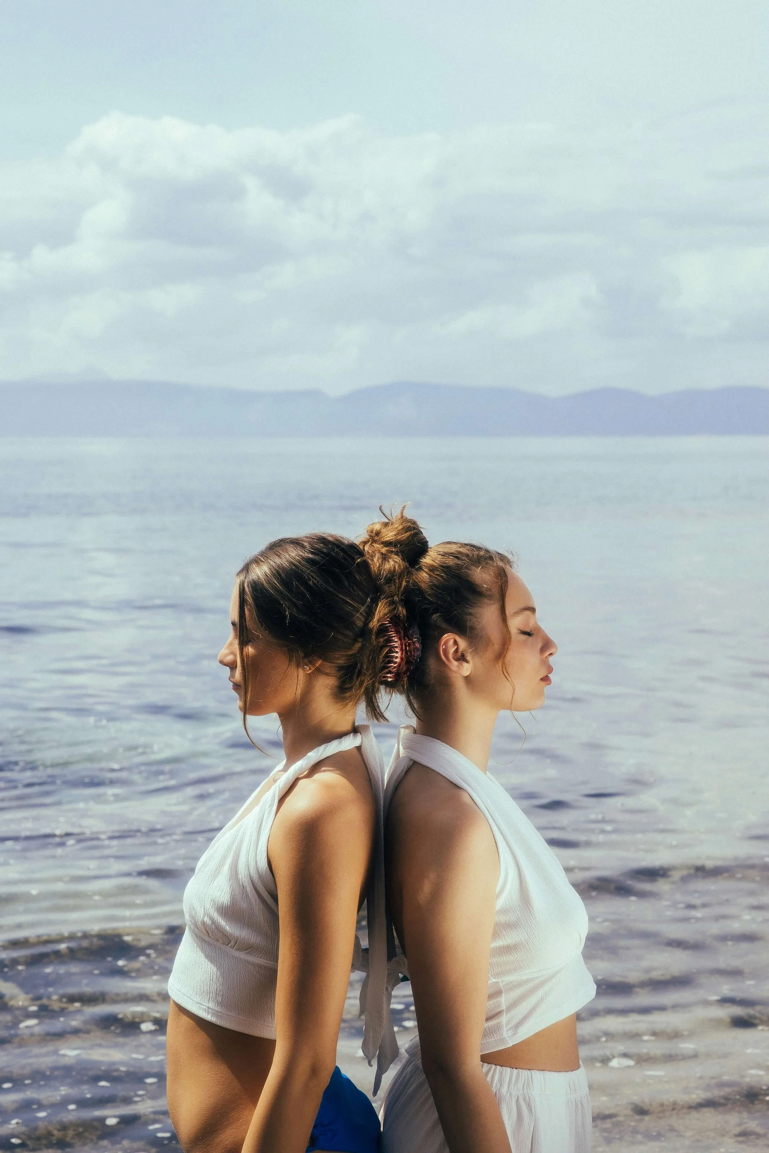 Two young women stand back-to-back on the beach near the water, with their eyes closed. They are wearing white sleeveless tops, and their hair is styled in loose buns. There are calm waters and distant mountains under a cloudy sky in the background.