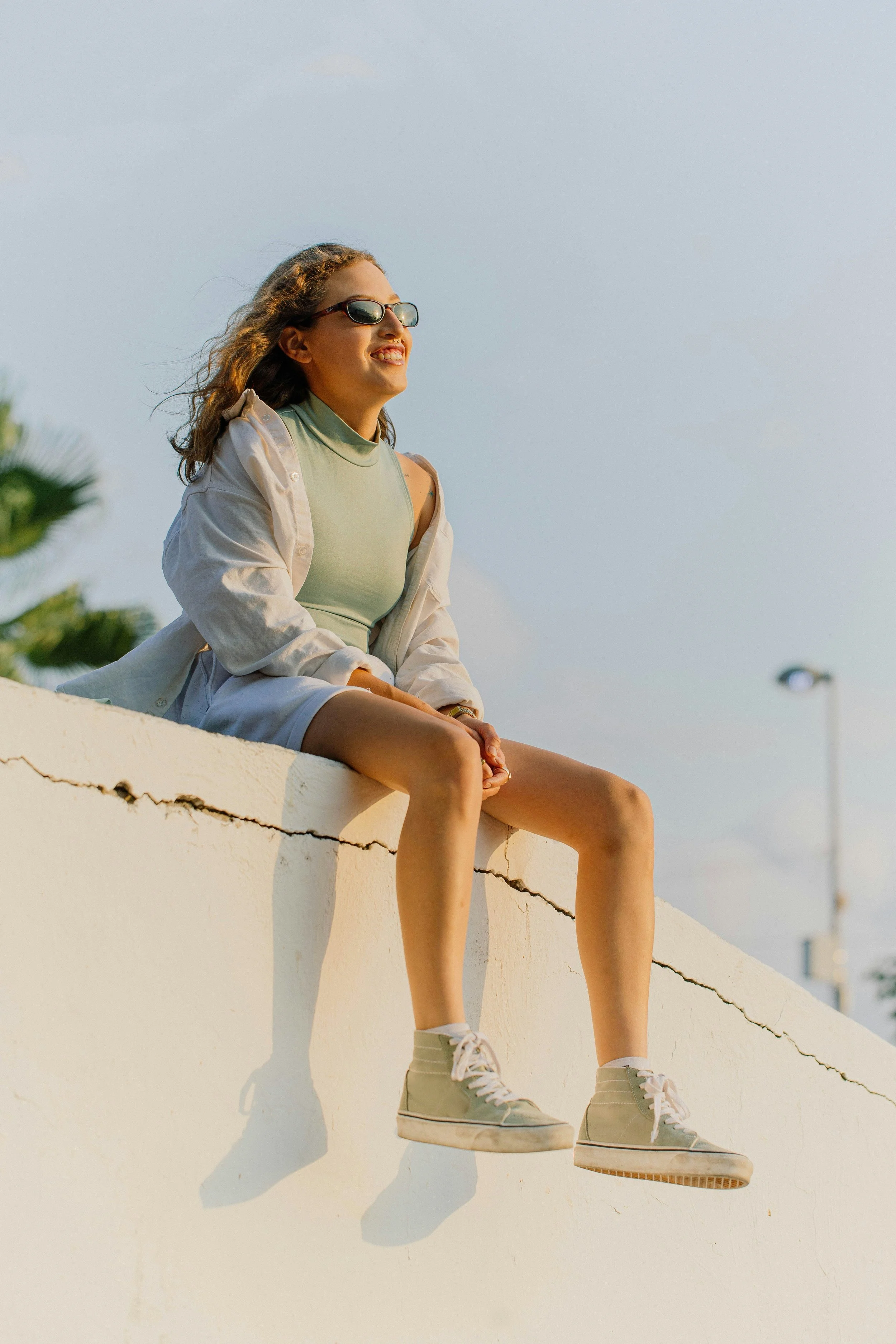 Young woman sitting on a white wall outdoors, smiling and looking into the distance, wearing sunglasses, a light green shirt, a beige jacket, shorts, and sneakers, with a partly cloudy sky in the background.