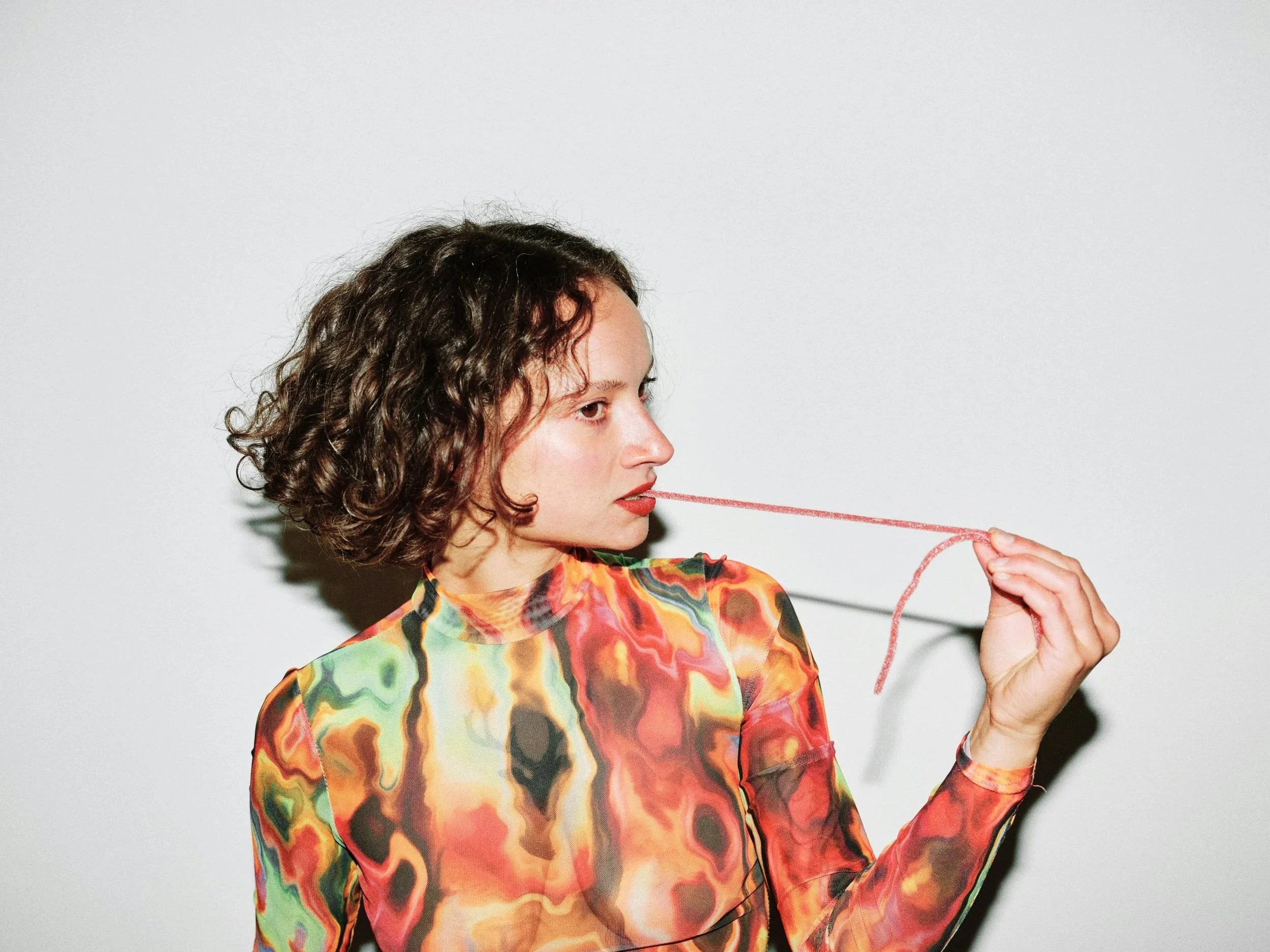 A woman with curly brown hair wearing a multicolored, abstract-patterned blouse or dress, holding a red yarn and looking to her right, in front of a plain light background.