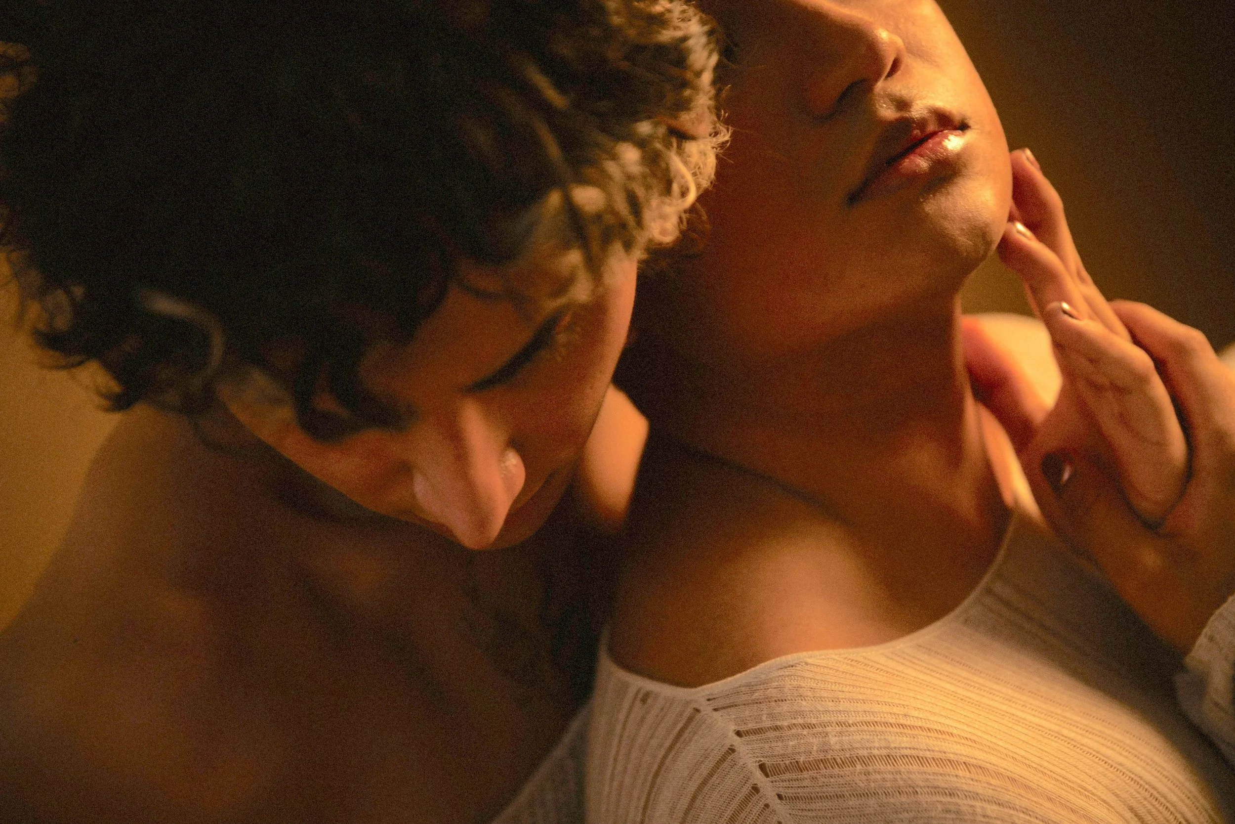 A close-up of two people in an intimate embrace, with one person's hand gently touching the other's face. The scene is softly lit, emphasizing emotional connection.