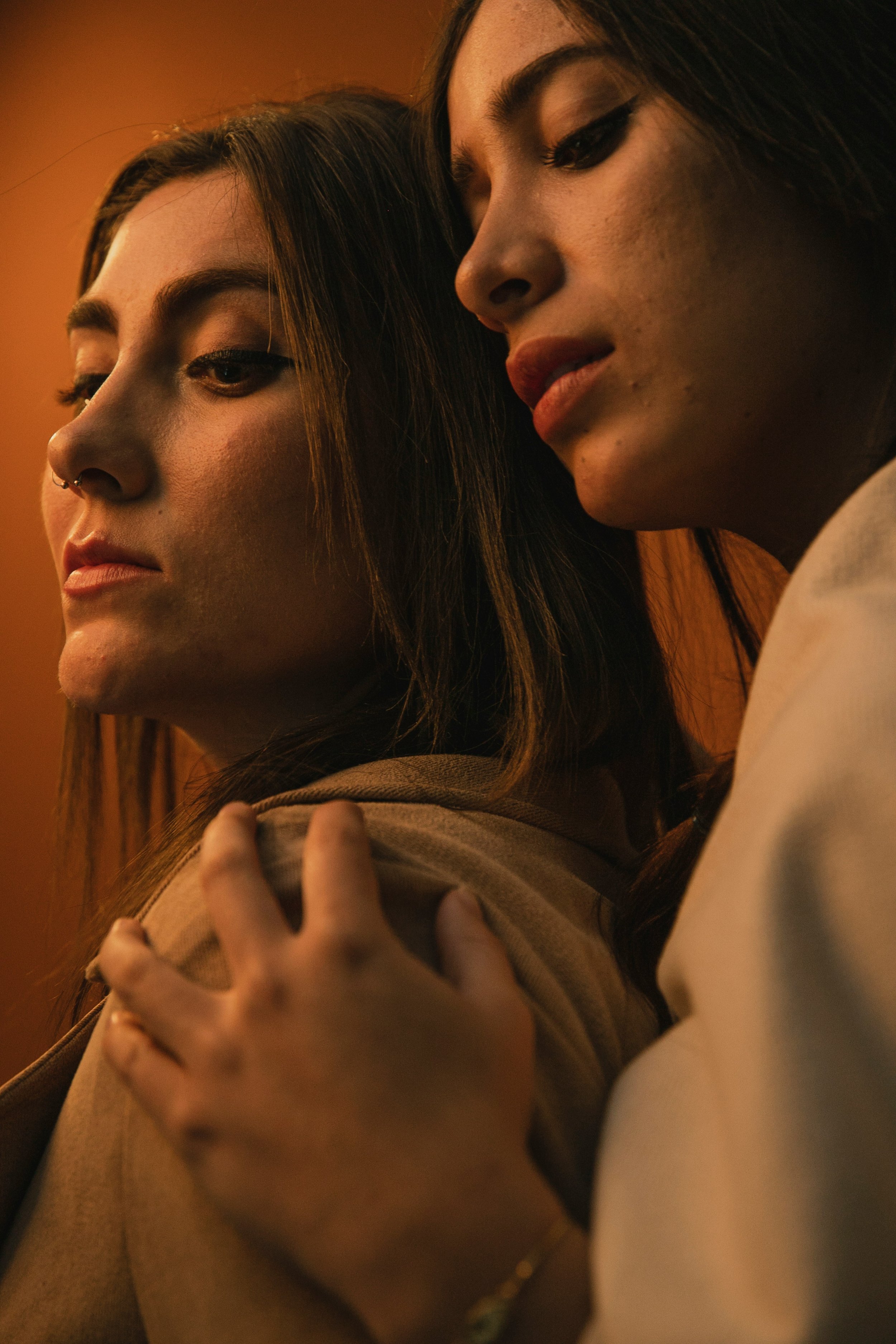 Close-up of two women with dark hair, one with winged eyeliner, quoting and looking away, in warm lighting.