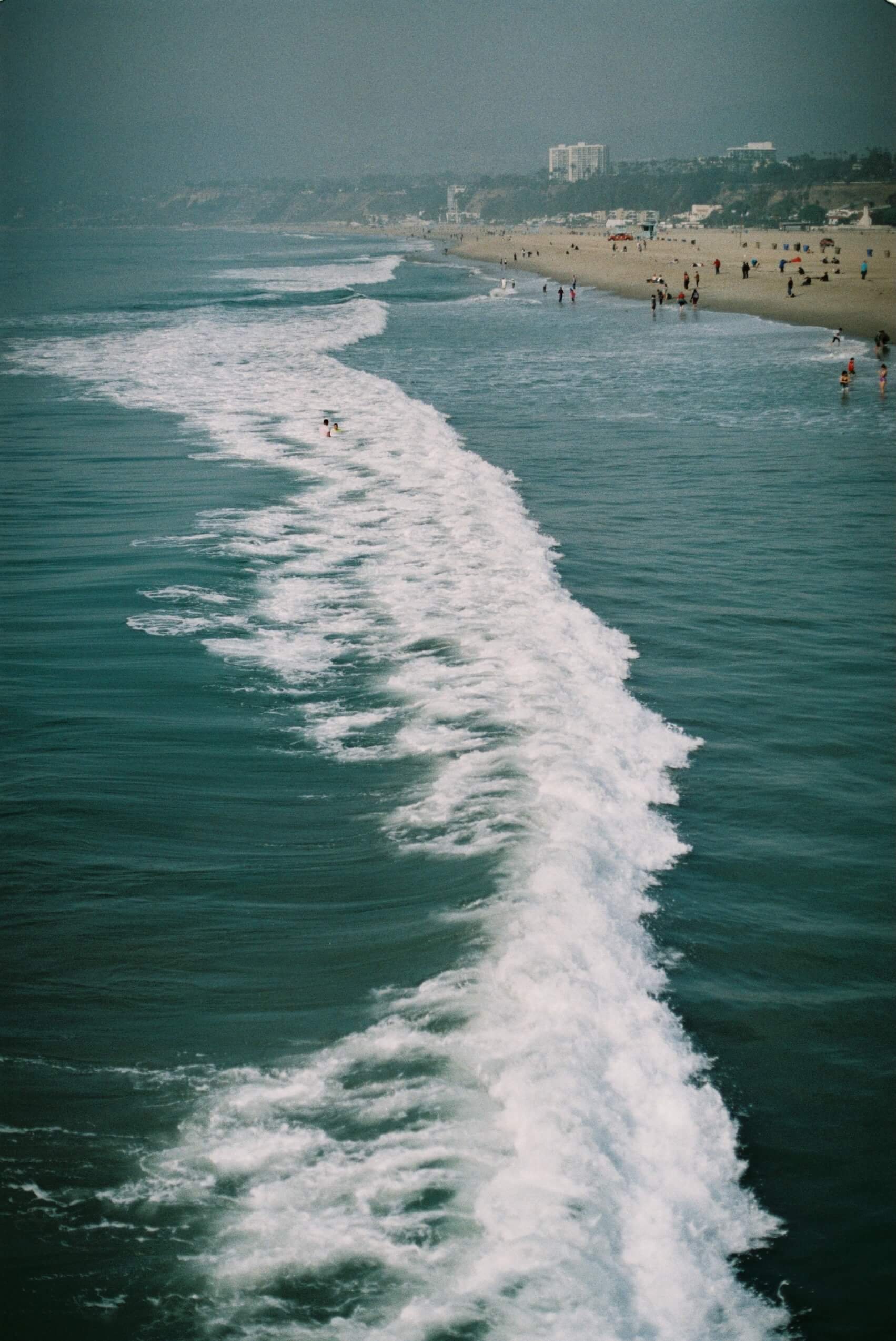 View from a boat showing the ocean waves in the foreground and a sandy beach with people in the distance, along with buildings and cliffs in the background.