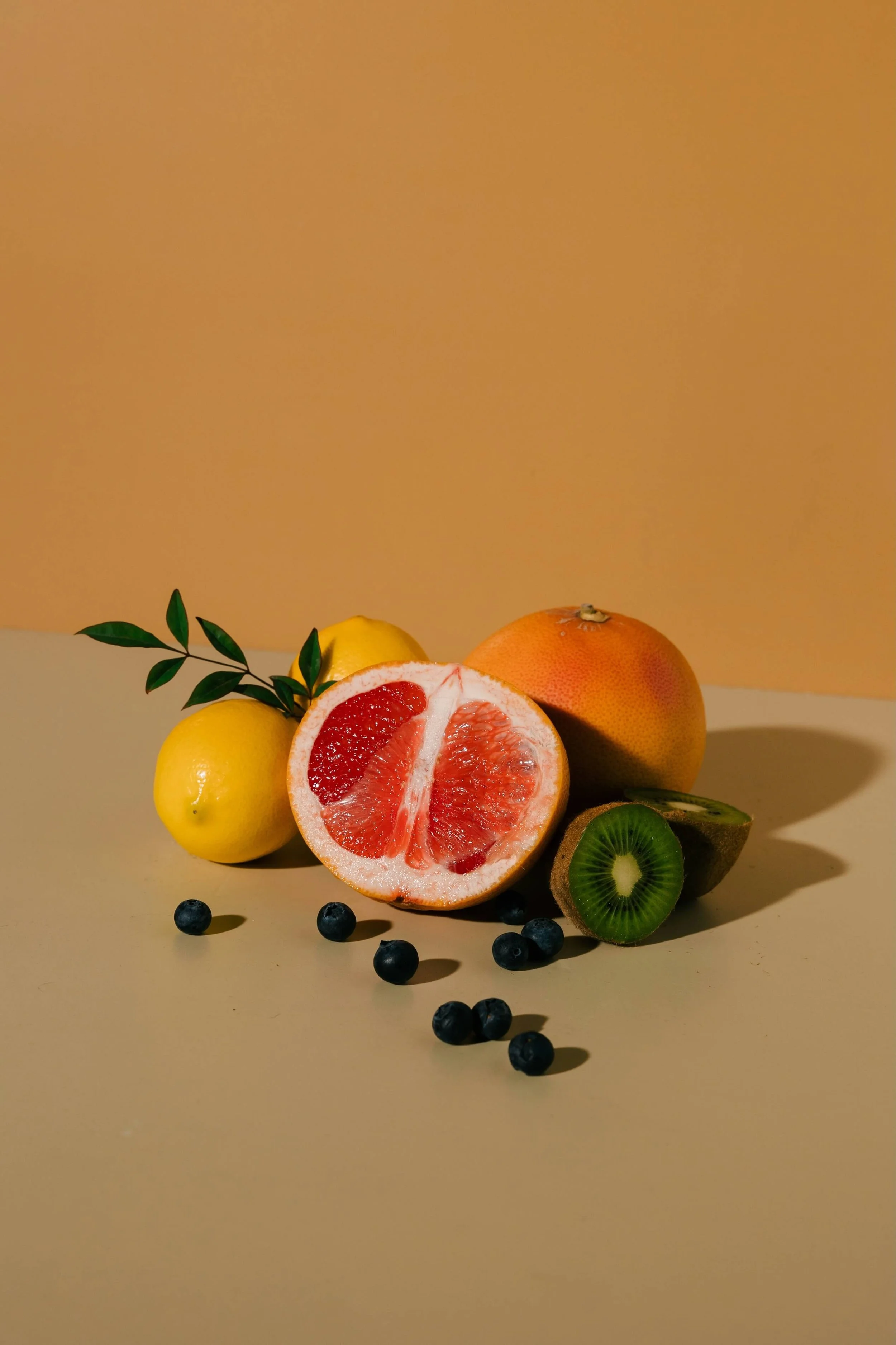 Assorted fresh fruit including lemon, grapefruit, orange, kiwi, blueberry, and a sprig of green leaves on a beige background.