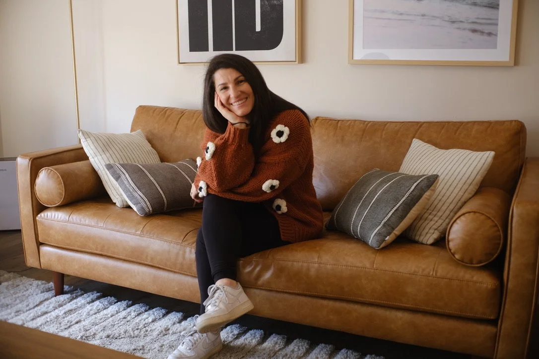 Woman sitting on a tan leather sofa with patterned pillows, smiling and resting her head on her hand in a cozy living room.