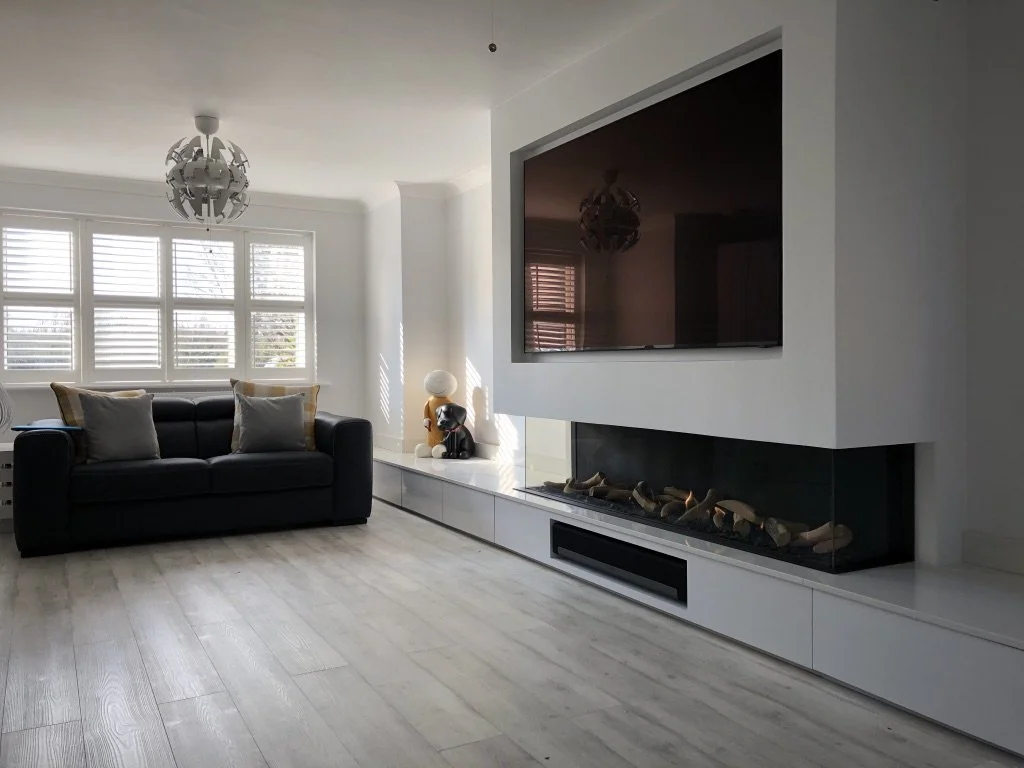 Modern living room with a black sofa, decorative pillows, a large wall-mounted flat-screen TV, a sleek fireplace beneath it, and a decorative lamp and figure on the white built-in fireplace mantle, with a window with white blinds in the background.