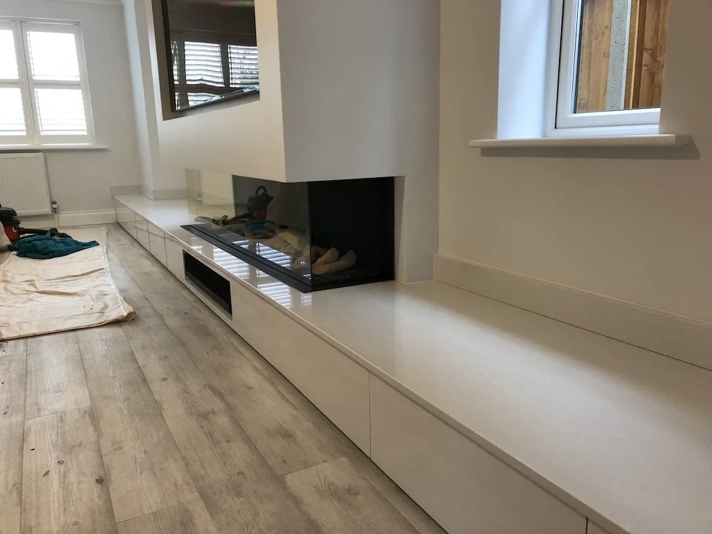 A modern white fireplace with a glass enclosure in a room with light wood flooring and white walls. There is a window on the right and a radiator on the left, with some tools and a cloth on the floor.