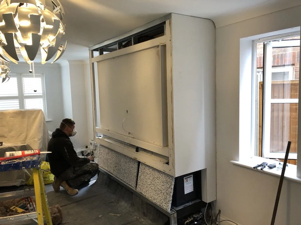 A person working on installation or renovation of a kitchen or living space, with a partially built structure and tools around, in a room with a window and modern lighting fixture.