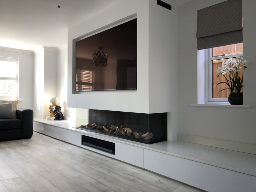 Living room with a wall-mounted flat-screen TV above a modern fireplace, black leather sofa, window with a gray shade, and a white orchid plant in a black pot.