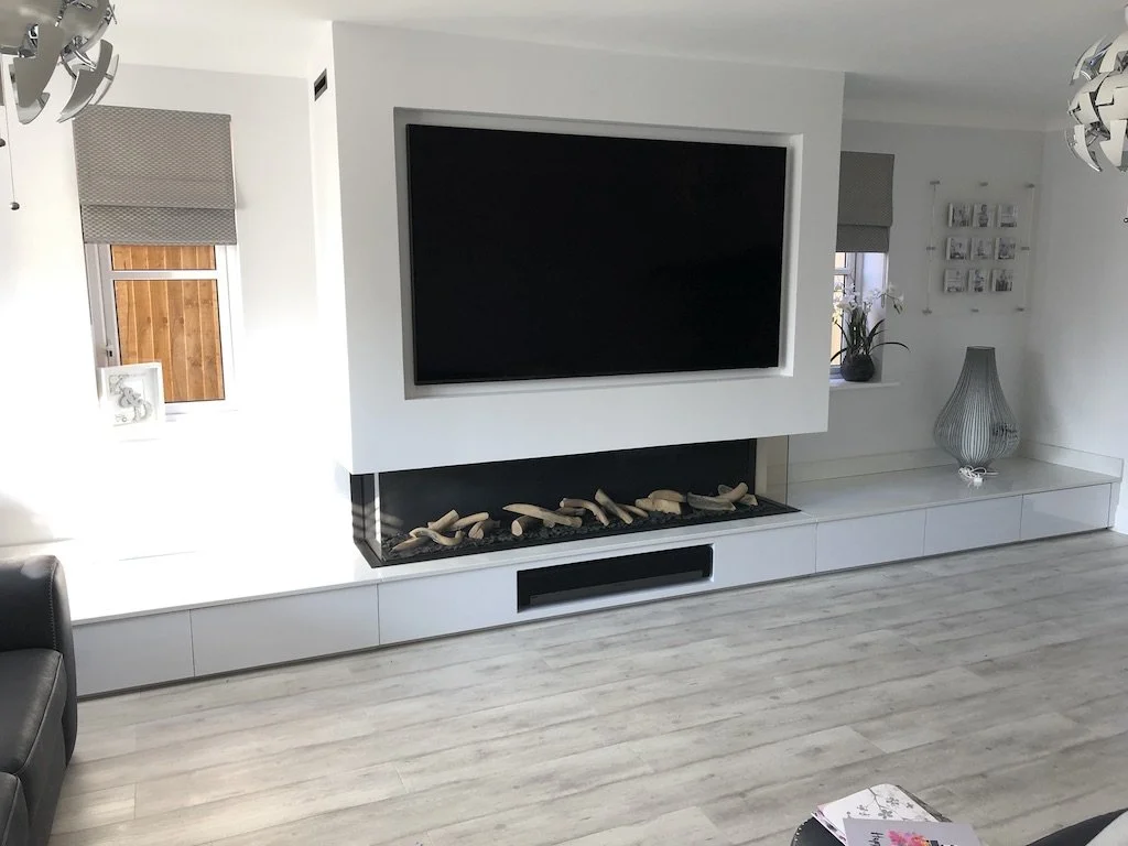 Modern living room with a large flat-screen TV mounted above a contemporary fireplace filled with decorative wood pieces. The room features light-colored wood floors, white walls, and decorative accents including a vase with plants, framed photos, an