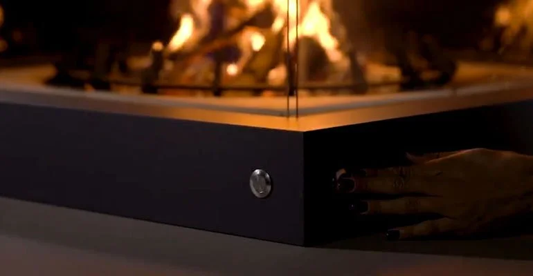 Close-up of a modern indoor fireplace with visible flames and a hand reaching towards the side of the firebox.