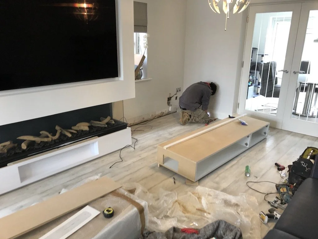A person installing a wooden media unit in a living room, with a black television and a modern fireplace on the wall, surrounded by tools and construction materials.
