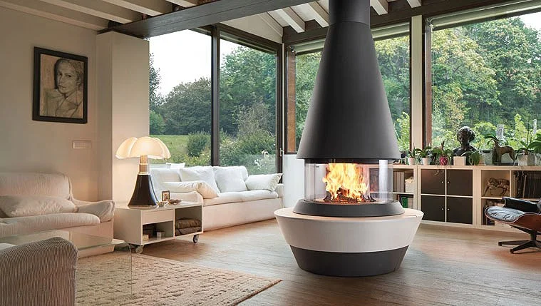 Modern living room featuring a central fireplace with a glass enclosure, large windows with outdoor greenery, a white sofa, a side table with a lamp, artwork on the wall, and various decorative plants and sculptures.
