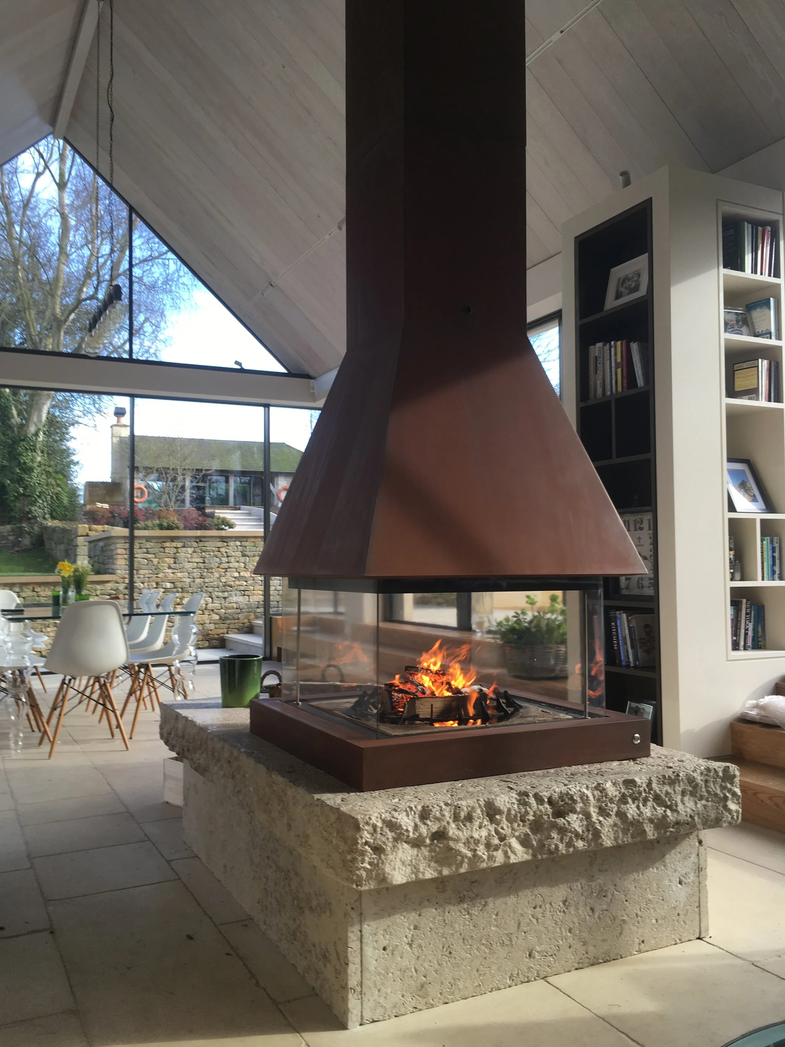Interior view of a modern living space with a central fireplace, a stone base, and a copper chimney. Large windows reveal a garden with trees, shrubs, and a patio with white chairs and a table. A white bookshelf filled with books and framed photos is visible to the right.