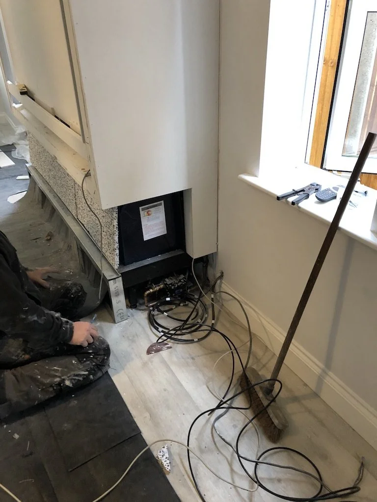 A construction worker installing a built-in fireplace with exposed wiring and scaffolding nearby in a room with a window.