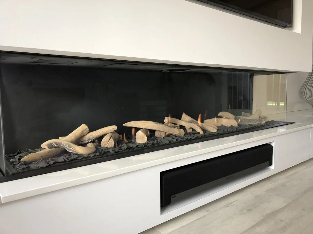 Modern built-in fireplace with white surround and glass front, containing decorative white firewood and small flames, located above a cabinet with a black vent.