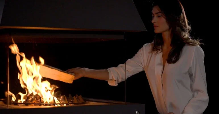 A woman in a white shirt tending to a fire in a fireplace or fire pit.