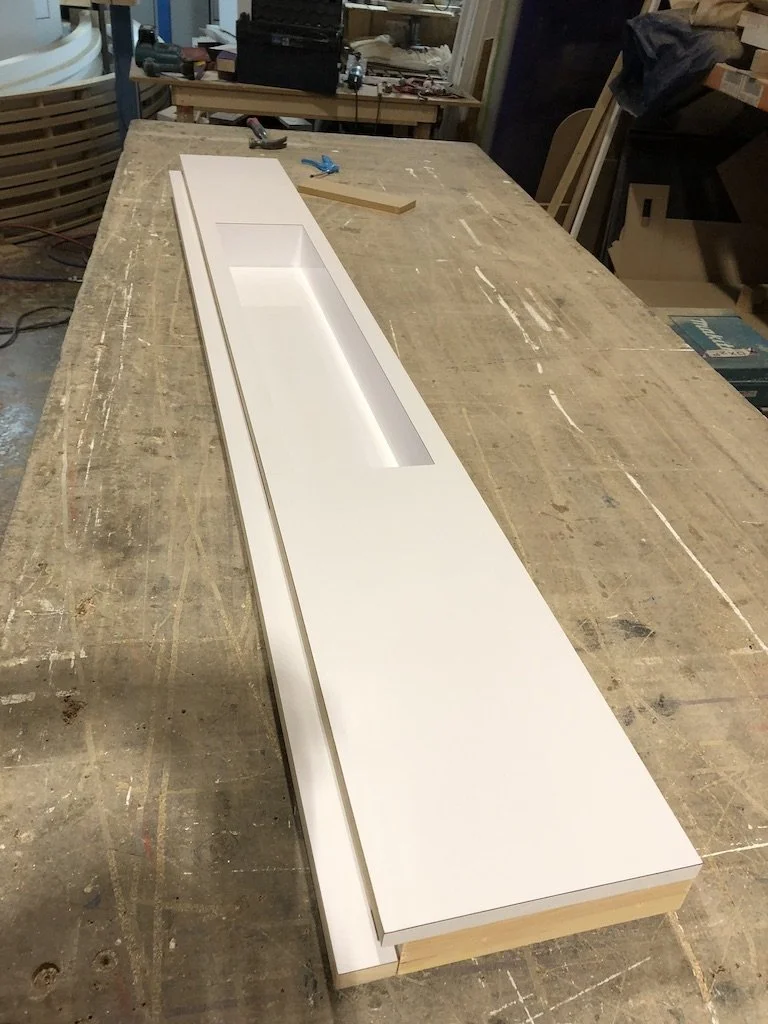 A long, white, rectangular built-in shelf with a cutout section is on a workbench in a woodworking shop.