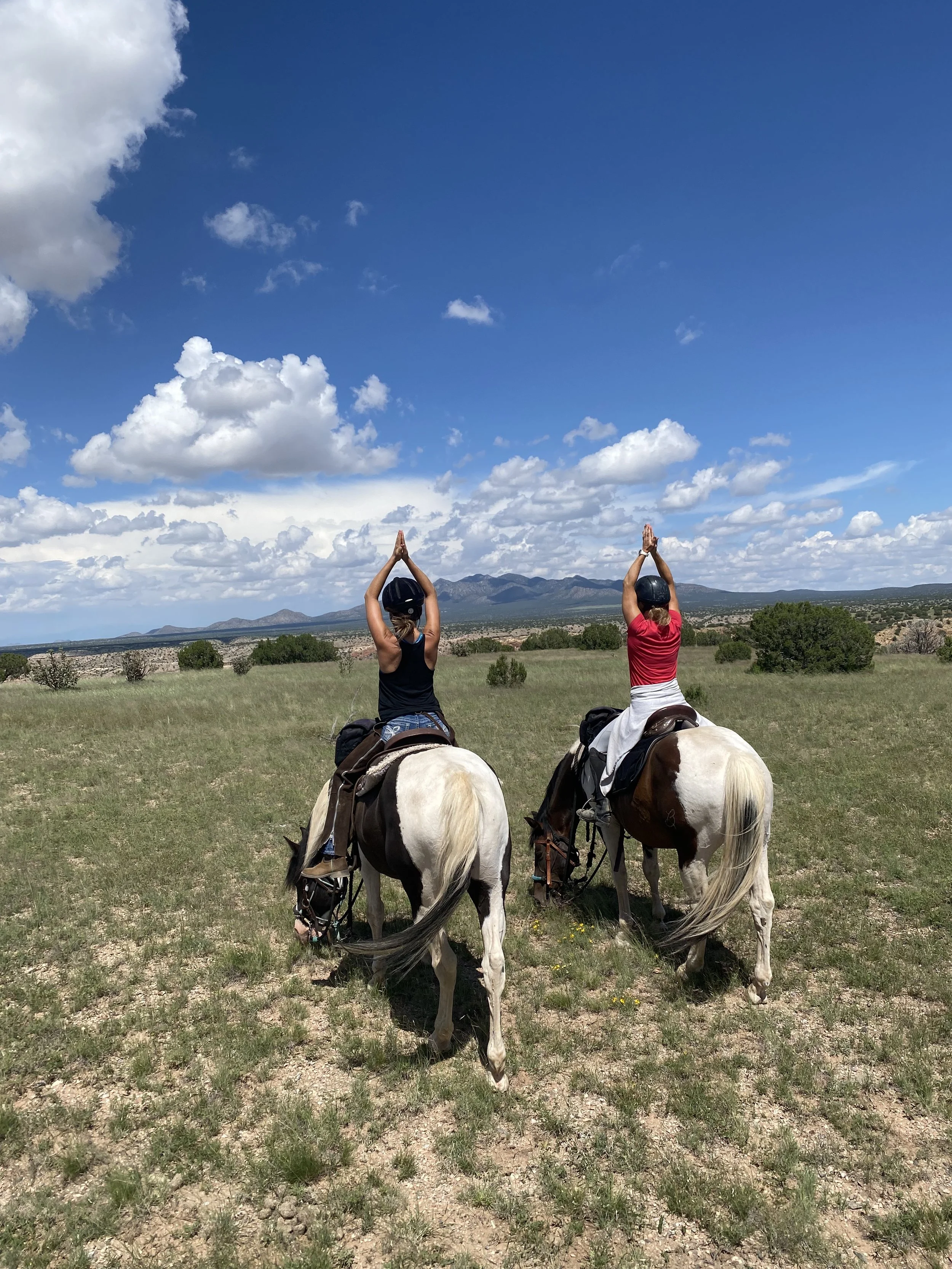 Two women riding horses in a grassy field with mountains in the background, under a partly cloudy sky. Both women are wearing helmets and have their hands raised in a prayer position.