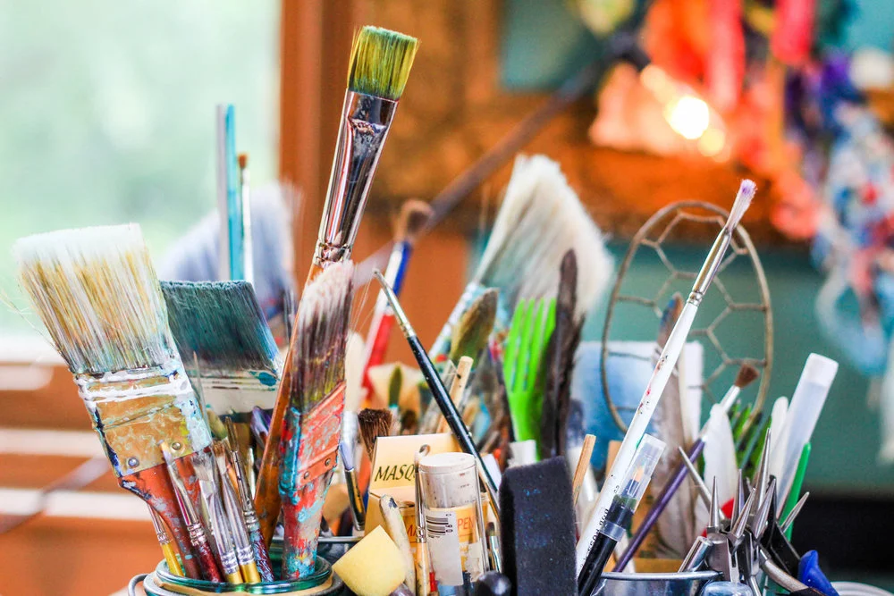A collection of various paintbrushes, markers, and art supplies in containers on a table, with a blurred colorful background.