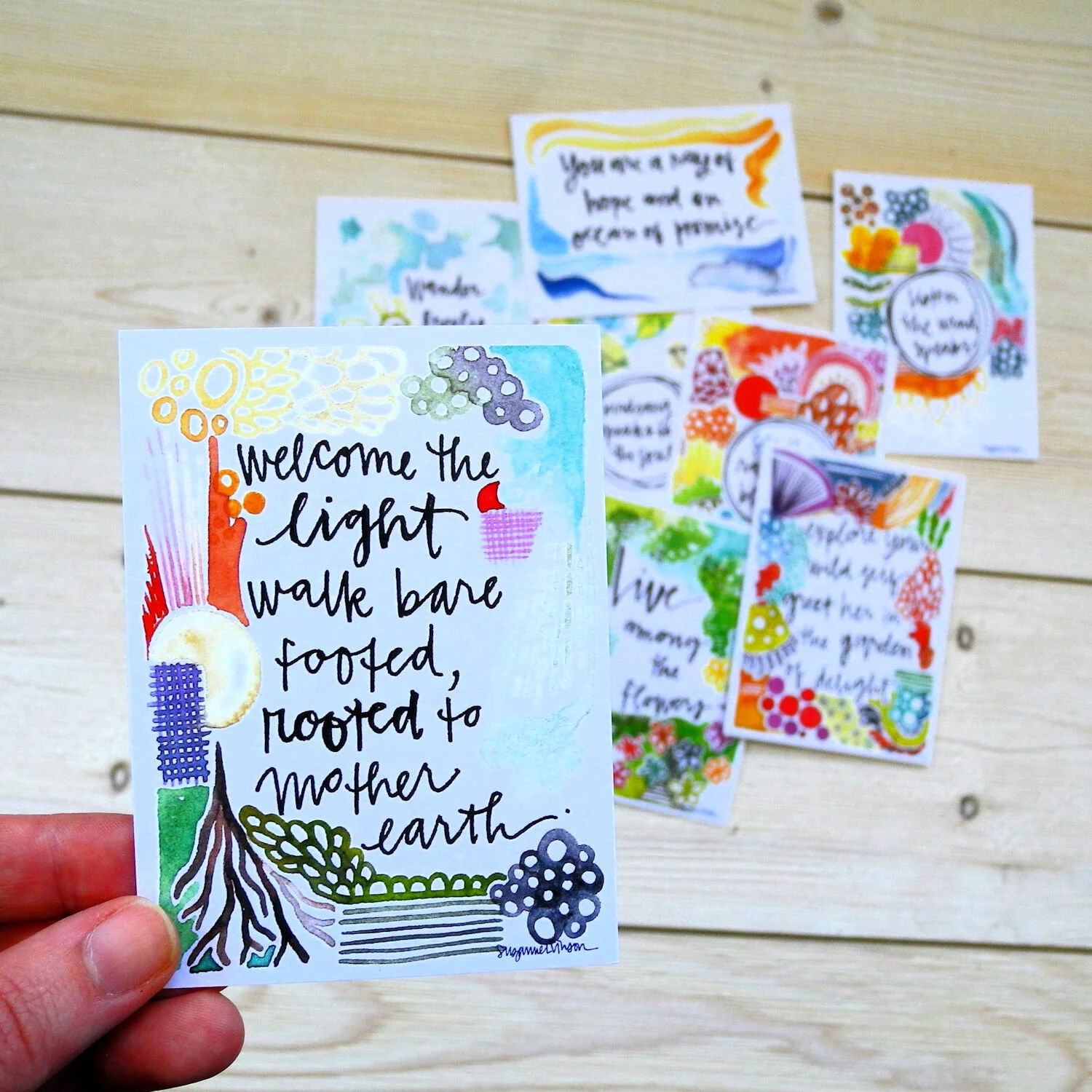 A close-up of a colorful hand-painted card with a quote about motherhood, featuring a tree with roots and vibrant flowers, held in front of several other similar cards against a wooden background.