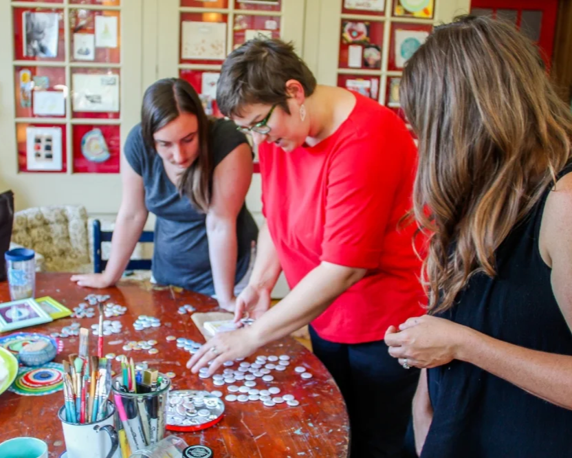 Three women standing around a table, playing a tabletop game involving chips and tokens, with a background of artwork on the wall.