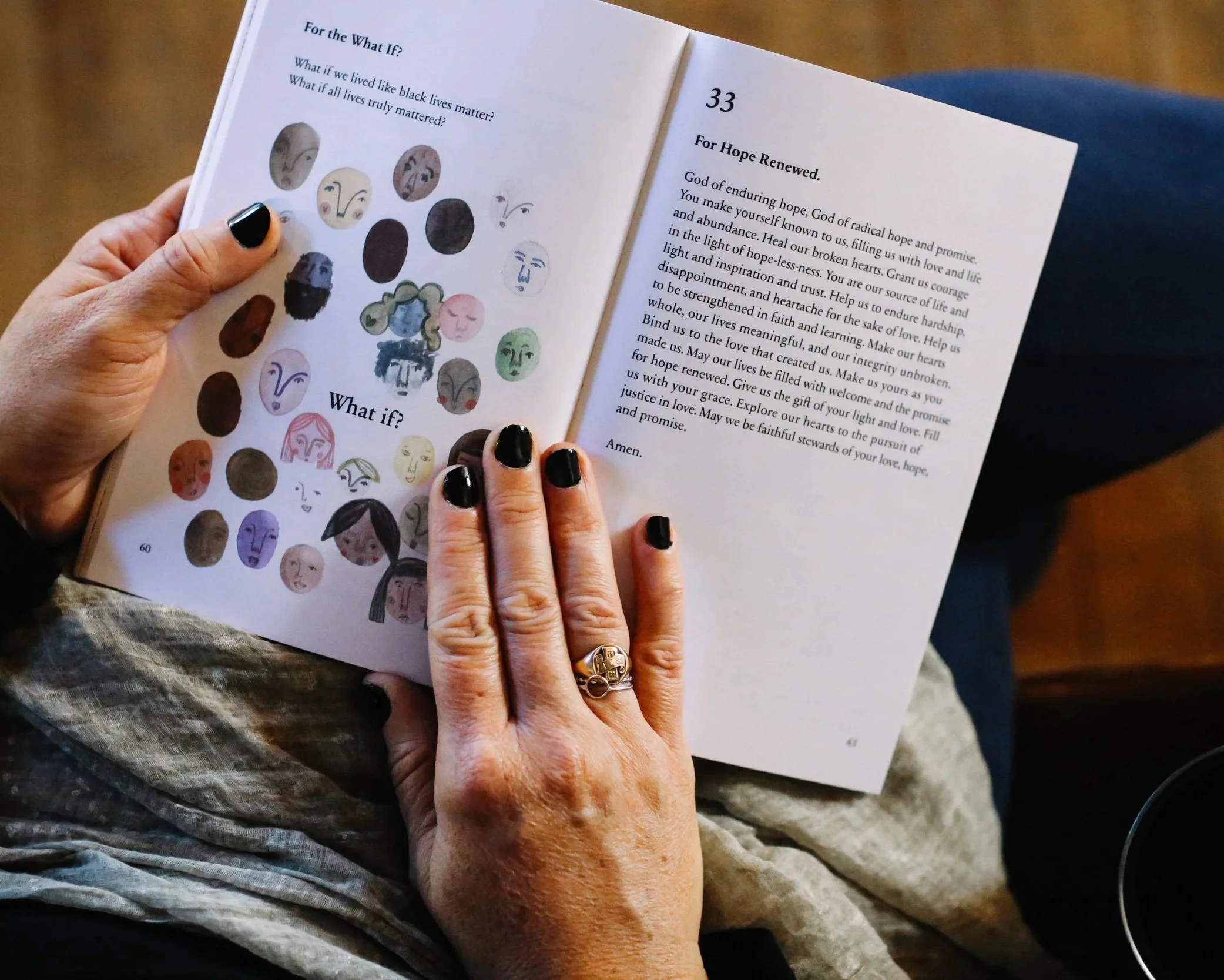 A person with painted nails holding open a book with the pages showing text and illustrations of various faces. The person's hands are visible, and they are wearing rings. The person's lap and part of their clothing are also visible.