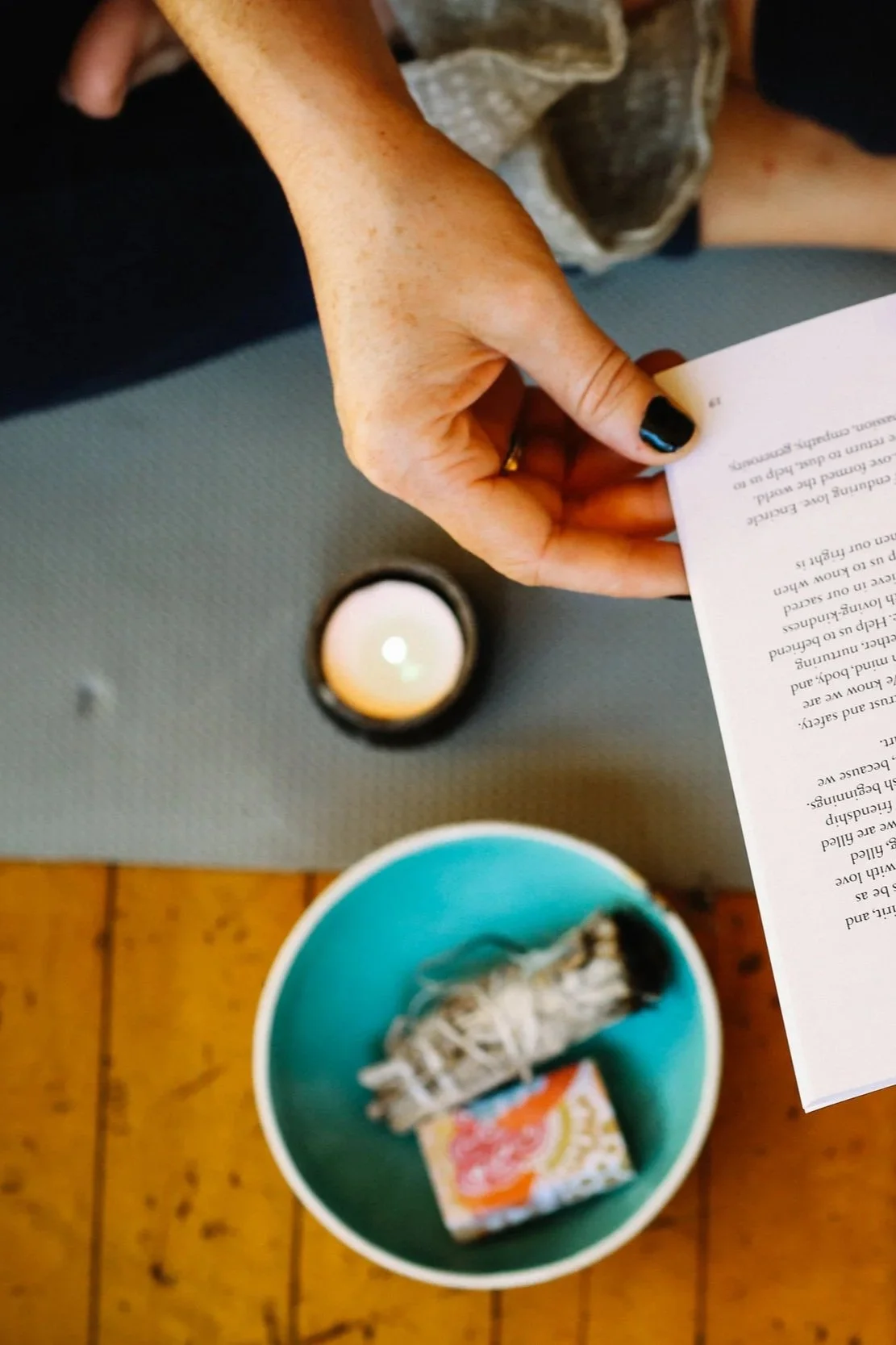 A person with a black-painted thumbnail holding a sheet of paper or a letter, sitting at a surface with a lit candle in a small container and a blue bowl containing a wrapped snack and a rolled-up piece of paper or napkin. The surface appears to be a gray mat, and the floor is wooden.