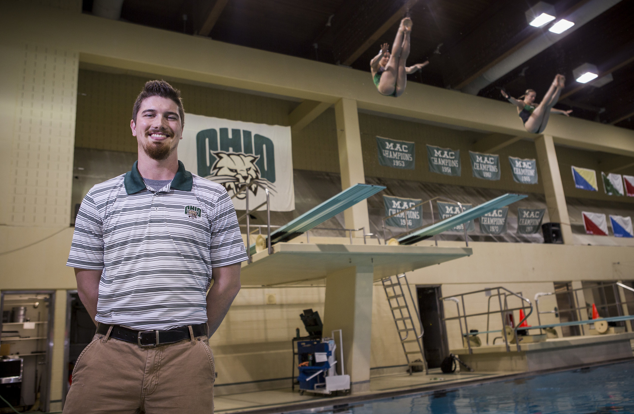  Tom Gimm, the new head diving coach, stands for a portrait as Ireland Littlejohn and Olivia Dillon dive in the background at the Ohio University Aquatic Center on Thursday, Jan. 26, 2017. 