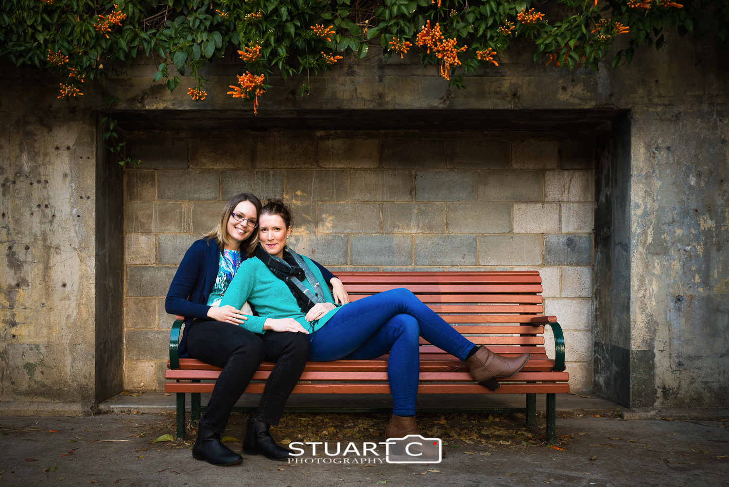female couple sitting on park bench in front of block wall with vines above