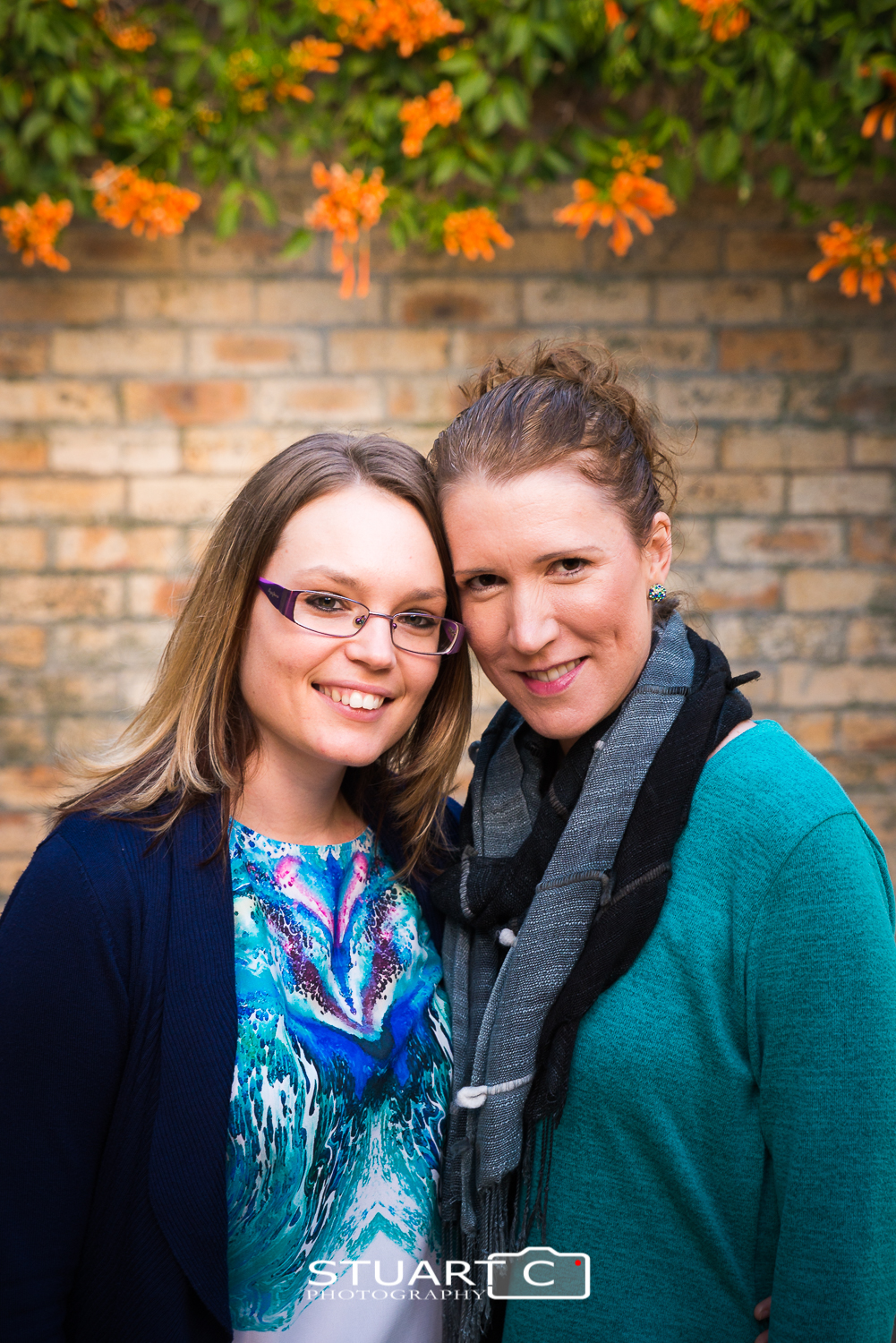 female couple in front of brick wall with orange flowers and green vine above