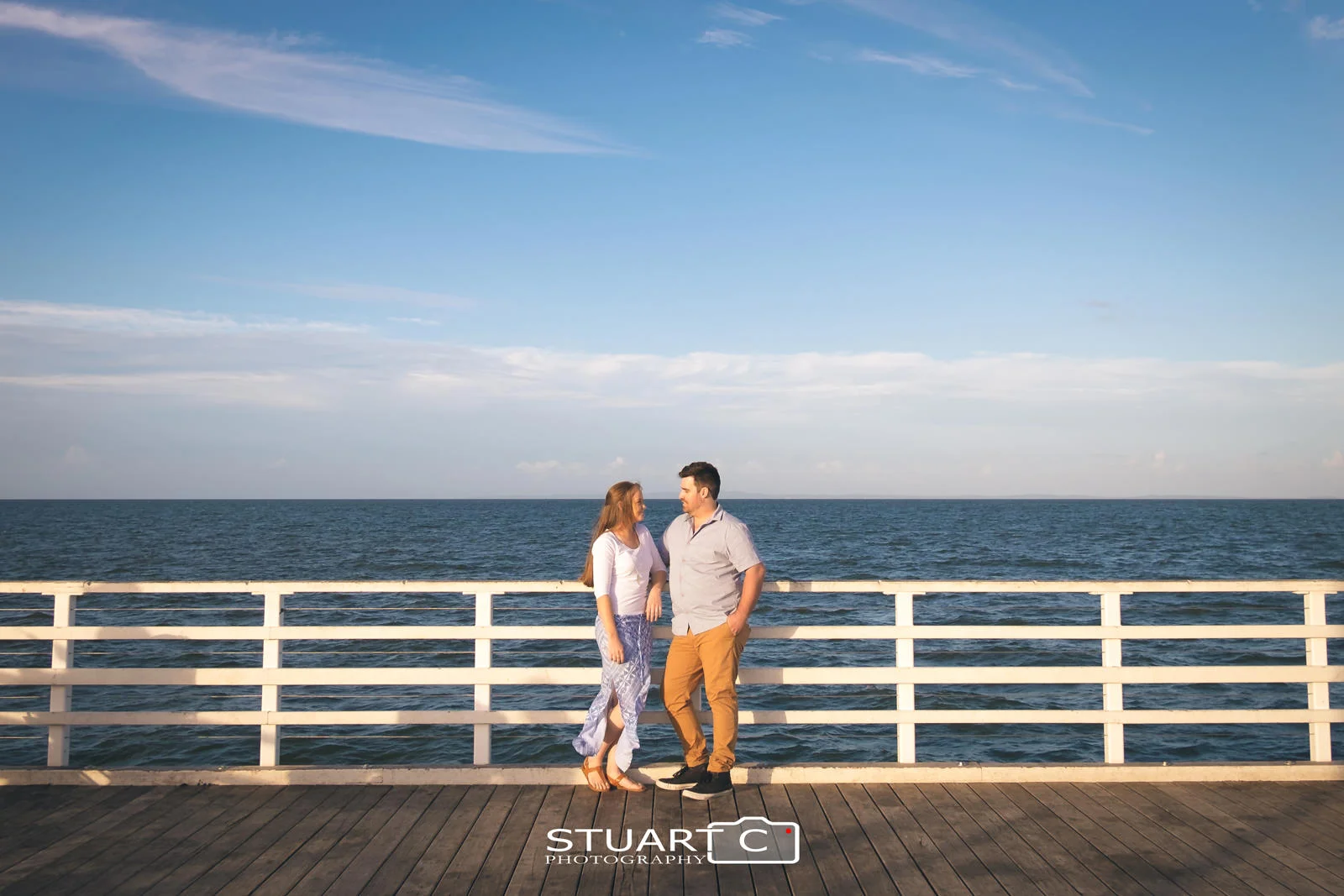 Wide environmental portrait of Engaged couple standing against white handrails at the end of Shorncliffe Pier with blue sky and clouds behind