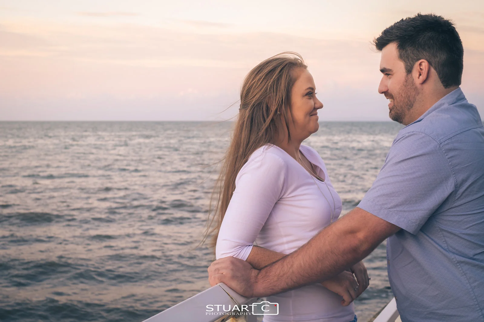 Portrait of engaged couple looking at each other standing against white handrail on shorncliffe pier during late afternoon sunset engagement photo shoot