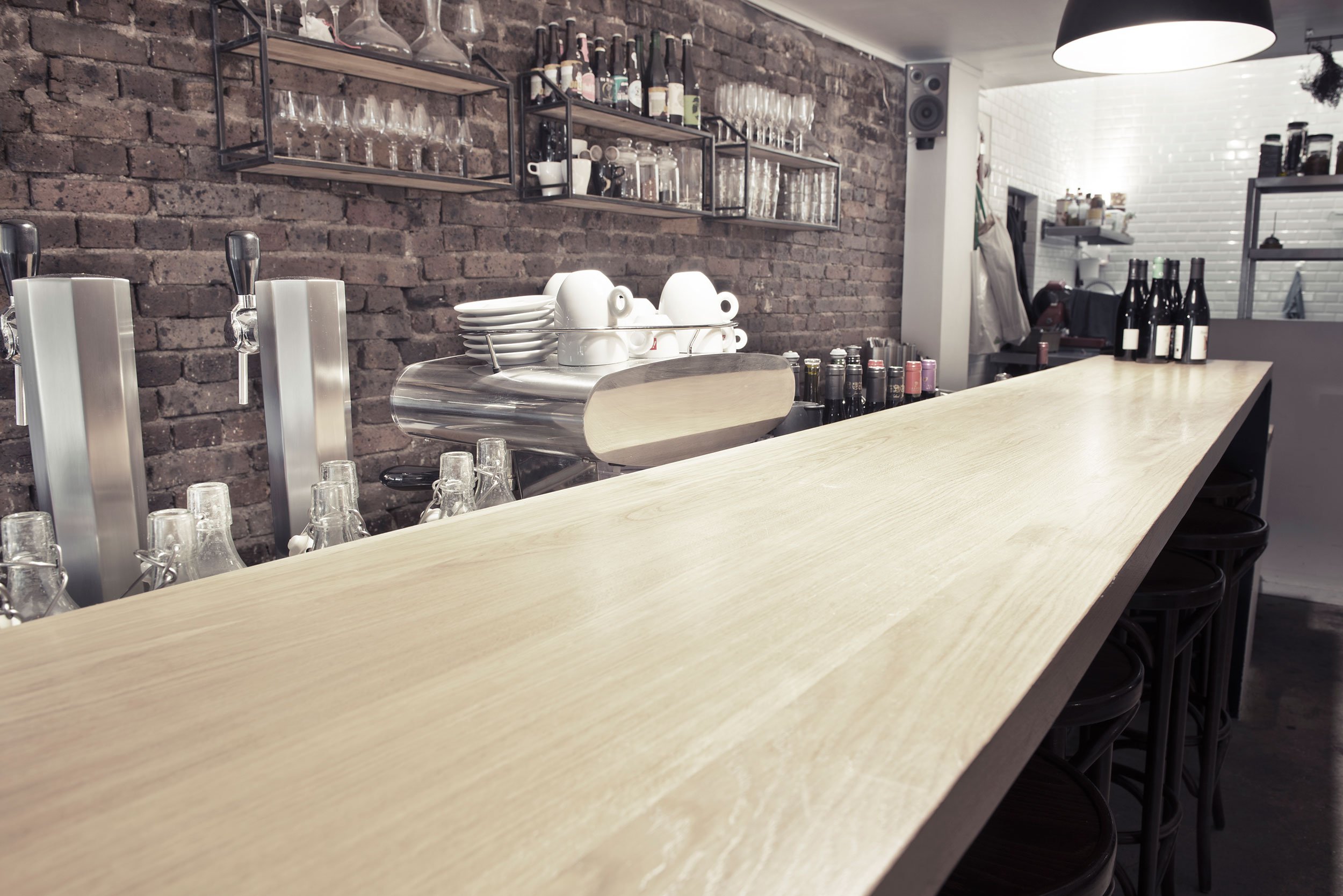 Comptoir du restaurant le Ballon Rouge, en bois clair avec verres à shooter, machine à café, tasses, bouteilles de vin, étagères en métal avec verres et bouteilles, murs en briques et en carrelage blanc dans un café ou bar moderne.