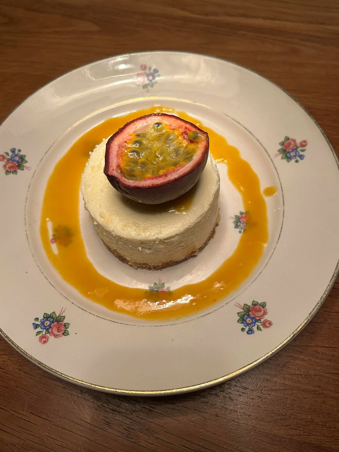 Gâteau au fromage avec une moitié de fruit de la passion sur le dessus, décoré d'une sauce à la mangue autour, servi sur une assiette à motif floral.