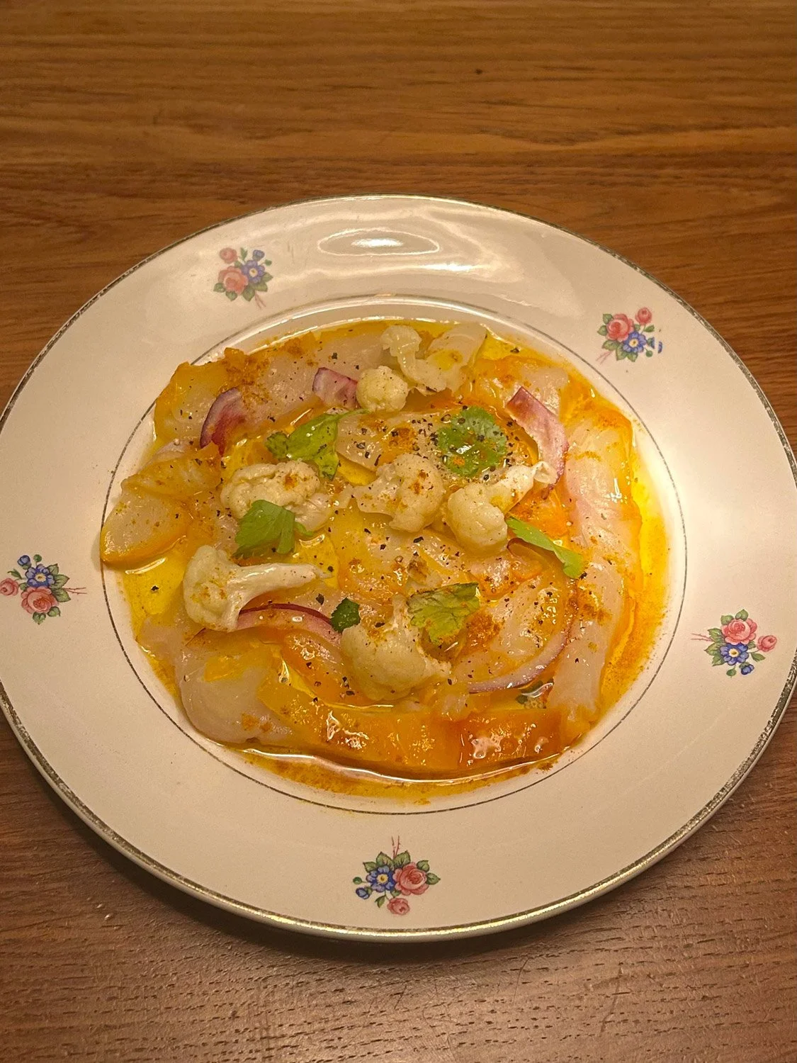 Menu du Ballon Rouge, poisson, légumes et épices dans une assiette à fleurs en porcelaine sur une table en bois.