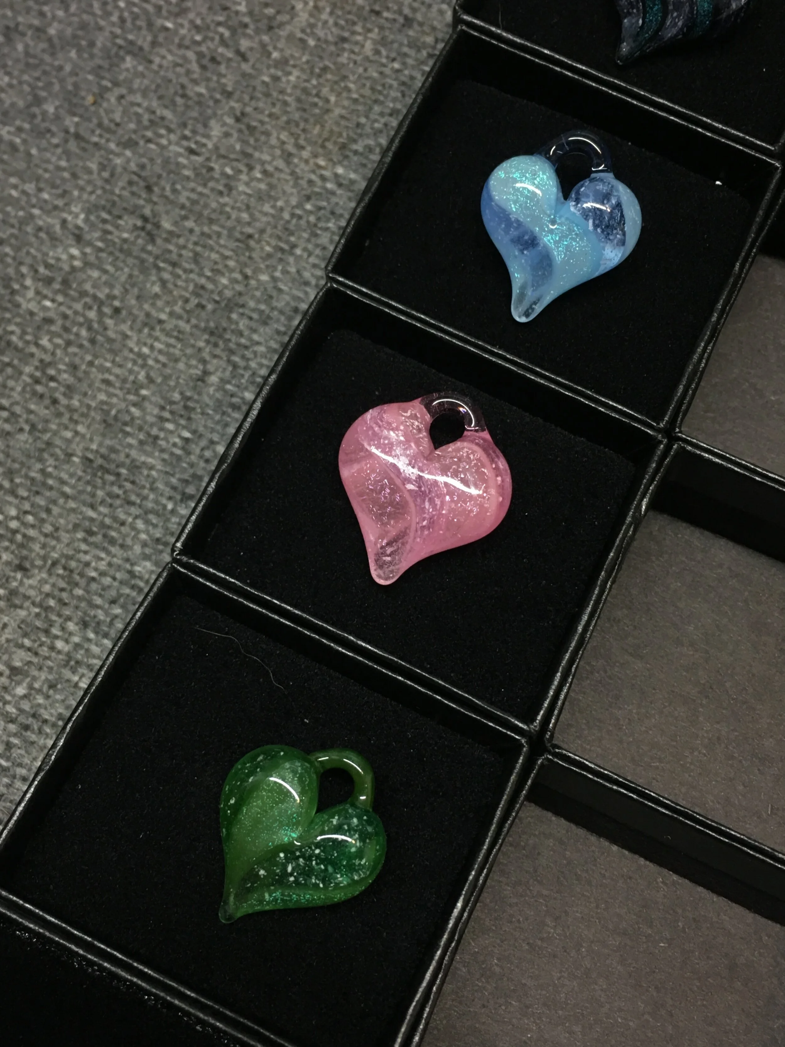 Three glass heart-shaped pendants in blue, pink, and green, each in its black display box on a black surface.