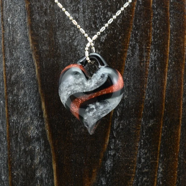 A heart-shaped glass pendant with black, red, and white swirl patterns hanging from a silver beaded chain against a dark wooden background.